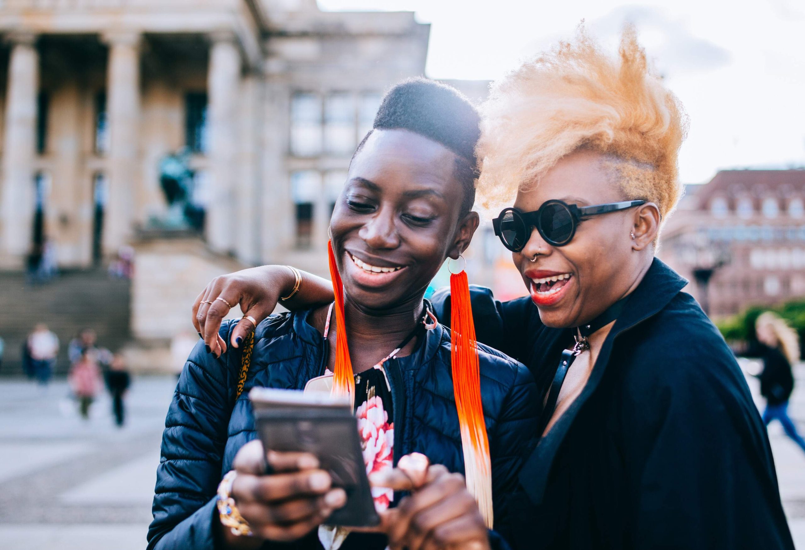 Two friends, one with orange hair and the other with long colourful earrings, smile as they look at a smartphone.