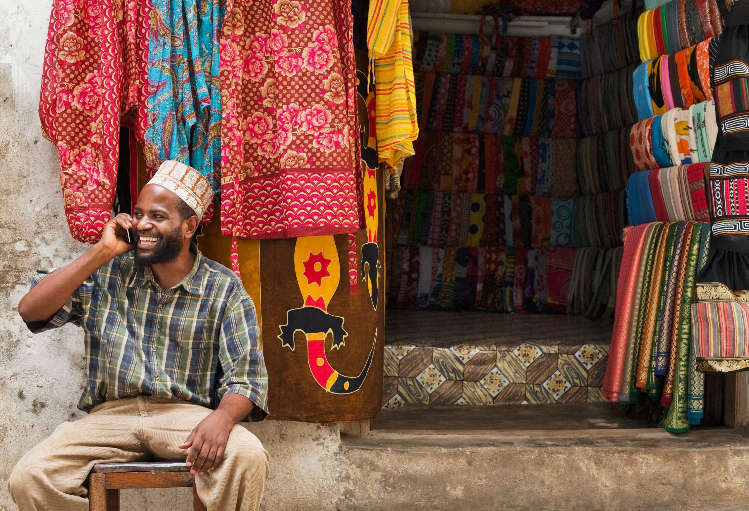 A cheerful man in a checkered polo engages in a lively phone conversation, seated outside a colourful drapery shop, as the vibrant patterns and textures form a captivating backdrop.