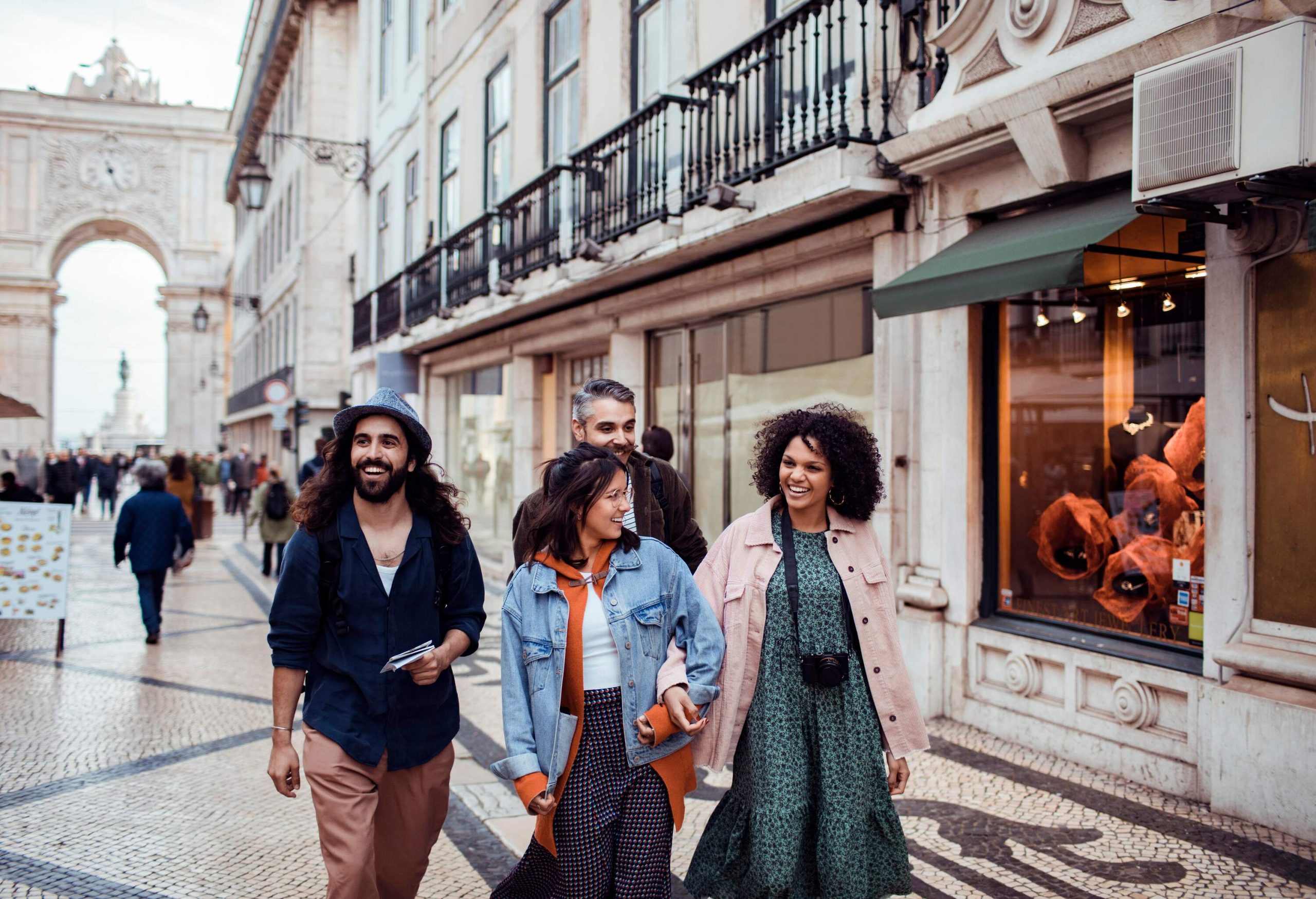 Four people walking across a cobbled market square, passing by quaint shops.