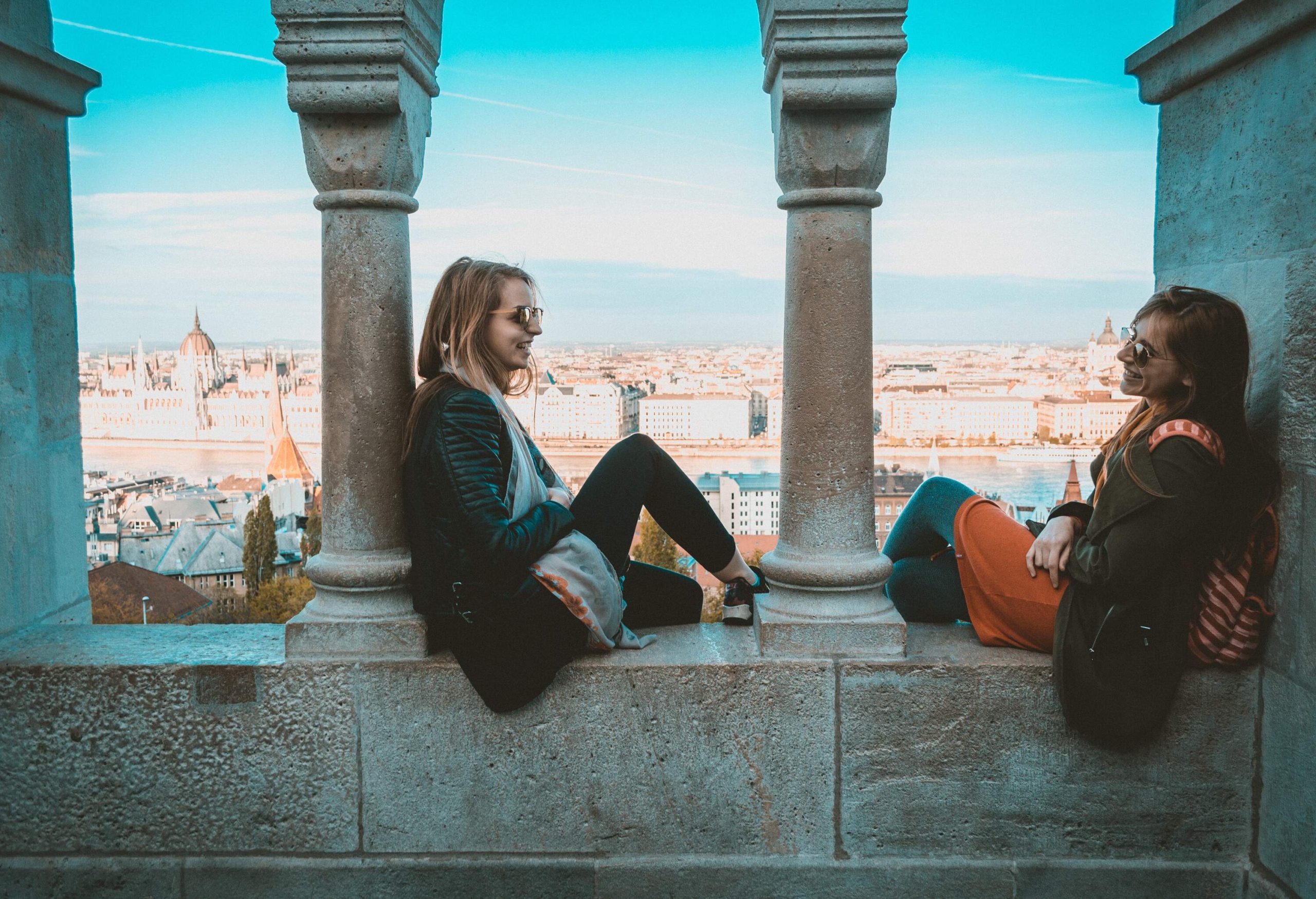 Two female friends talk while sitting on a pillared balcony's ledge.