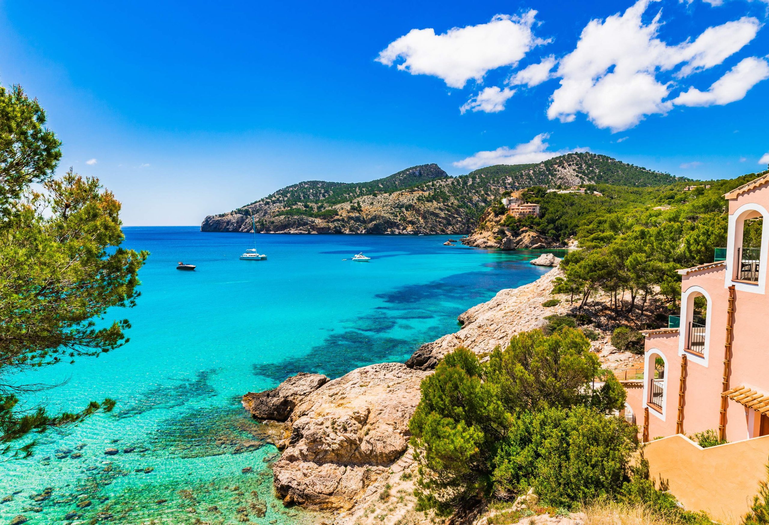 A calm blue sea surrounded by lush green rocky mountains, blue skies and white clouds.