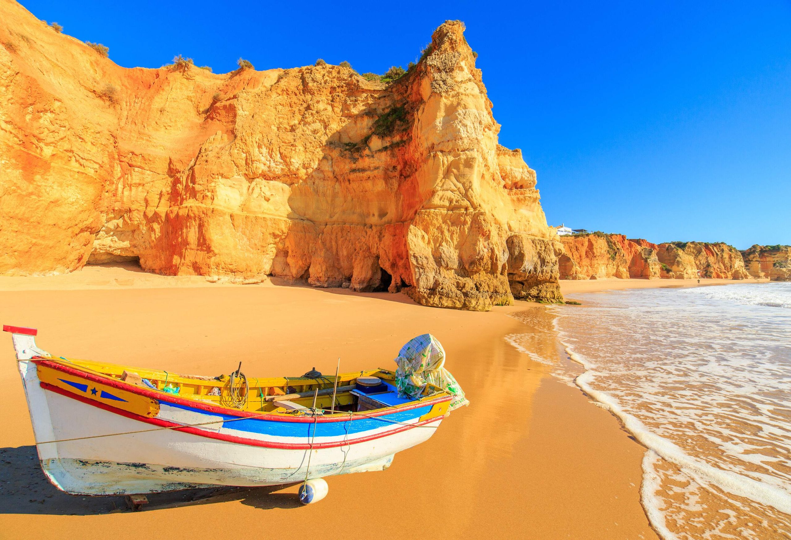 A colourful fishing boat on the shore of a fine sandy beach surrounded by tall rugged cliffs.