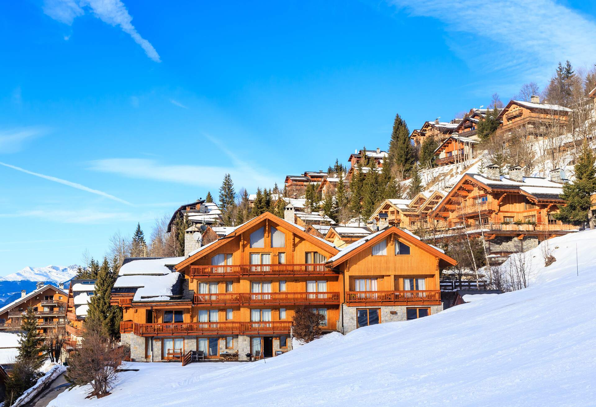 Wooden chalets along the slope side of a snow-covered mountain dotted with trees.