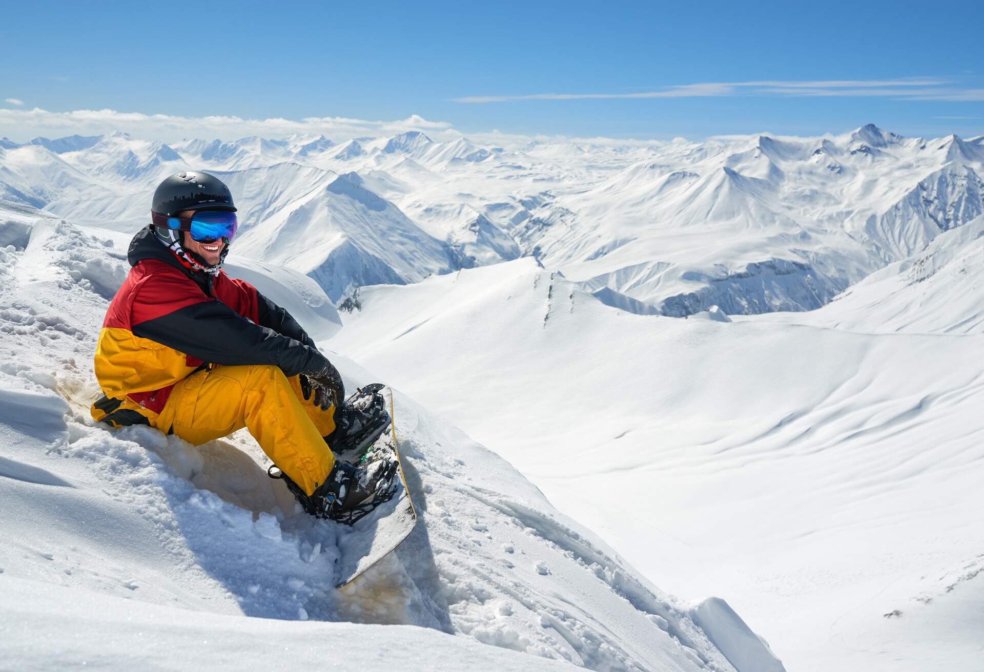 A man with a snowboard on sitting on a slope and gazing out into the snowy landscape.