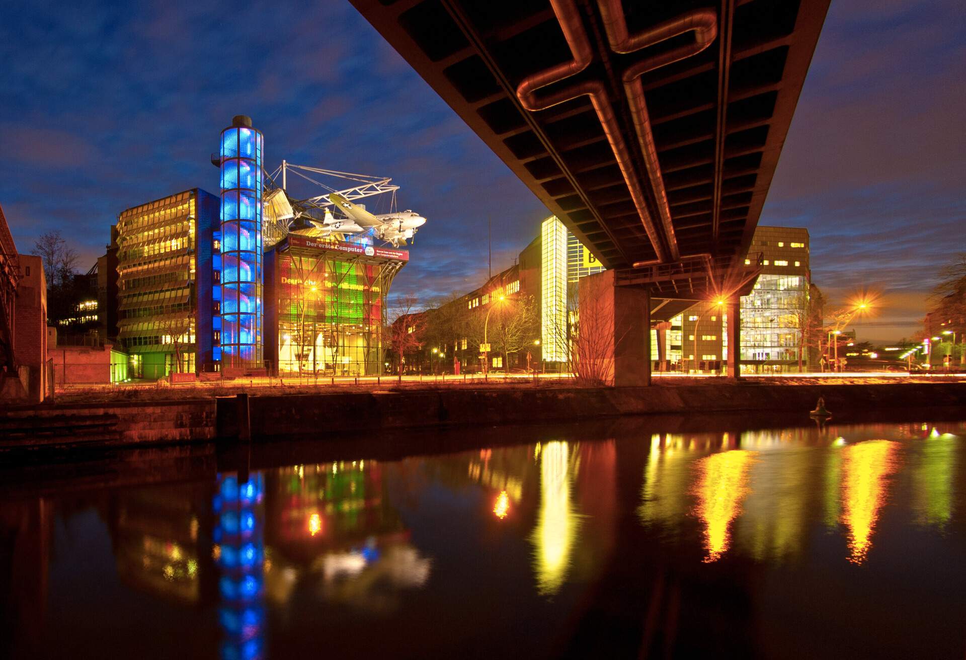 Brightly lit museums with transparent glass walls reflect in the waters seen from beneath the bridge.