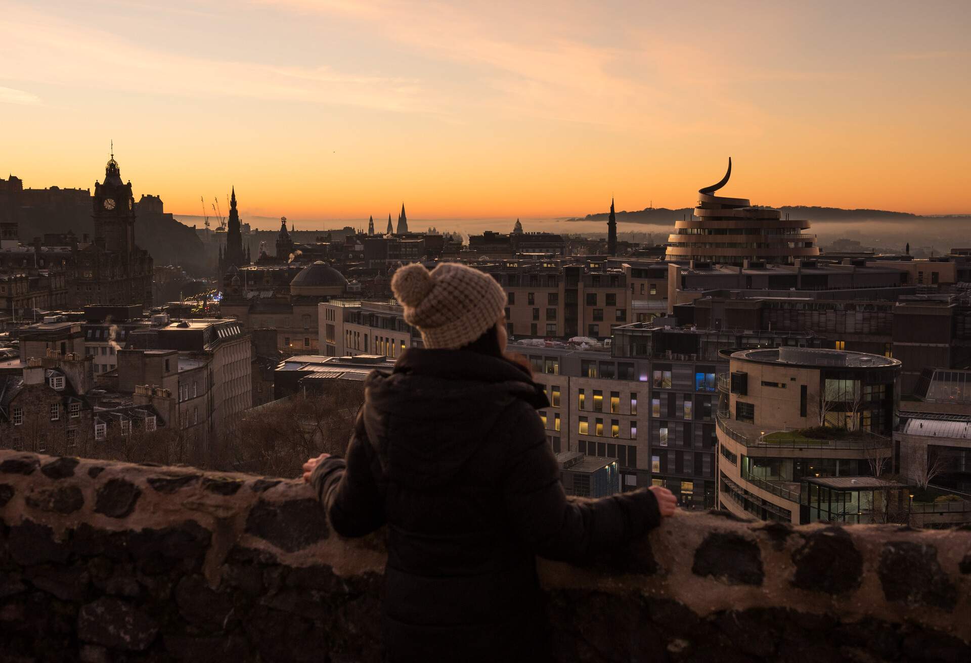 Asian woman wearing a winter jacket and a woolly hat admires the view of Edinburgh City's skyline during a misty sunset with a dramatic red sky and low clouds in Scotland, United Kingdom
