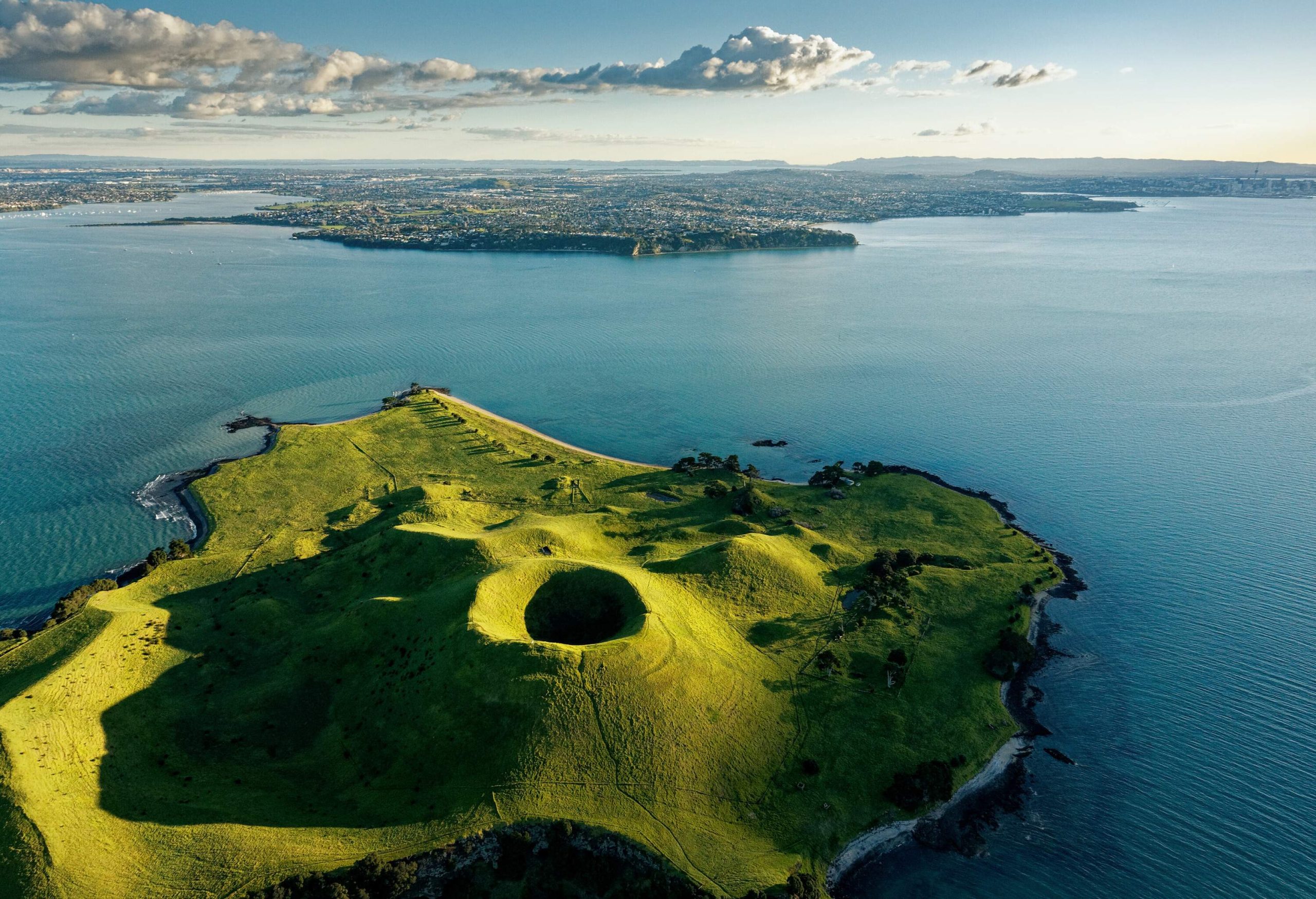 A spectacular sunlit conical volcano with a deep crater covered by lush green grass emerges on the sea surface.
