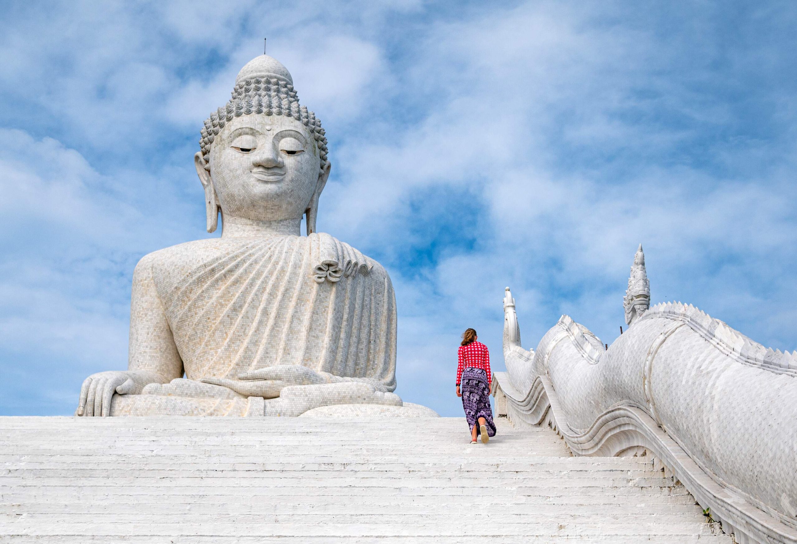 A young lady goes up the stairs to the Phuket Big Buddha statue made of white marble against the clouds in the sky.