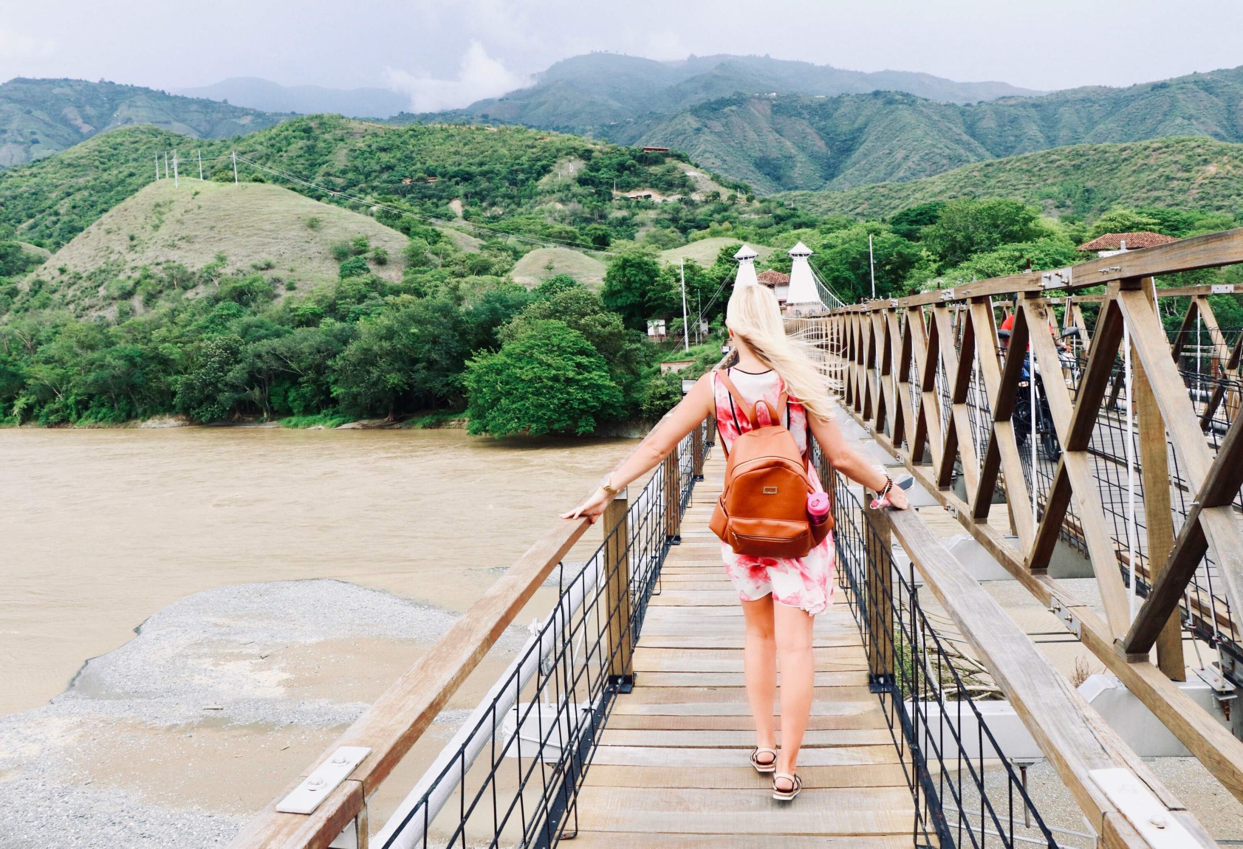 A blonde girl with a brown bag crosses a bridge across a river towards a mountainous area.