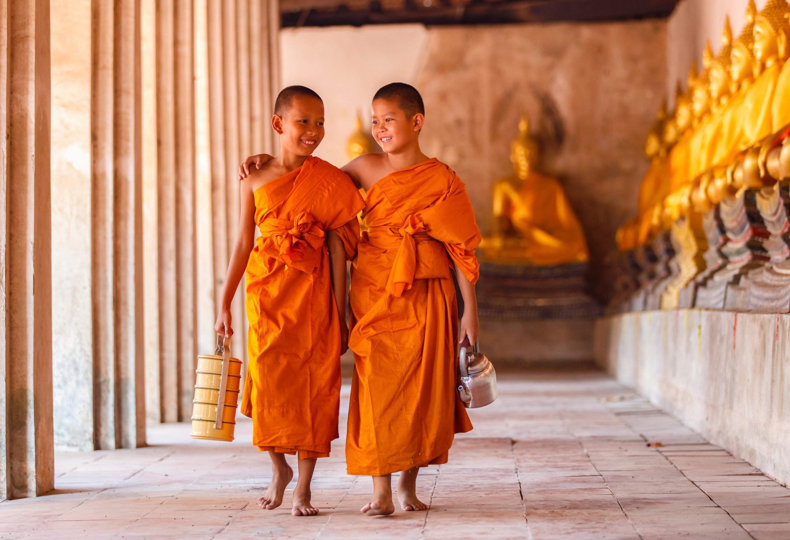 Two young monks in orange robes stroll along a temple hallway with golden Buddha statues.