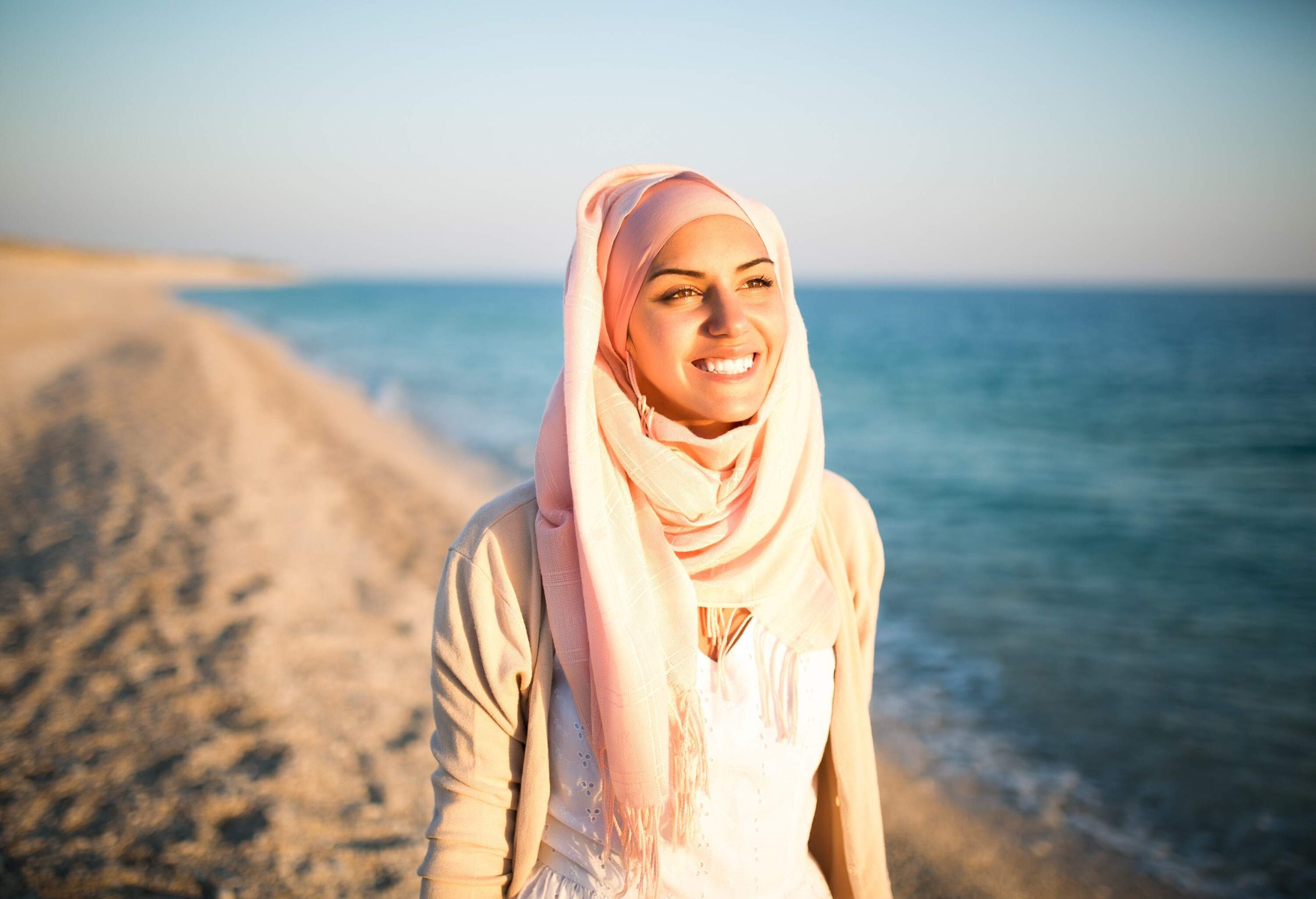 A happy woman in a pink hijab enjoys the sun by the shore of the beach.