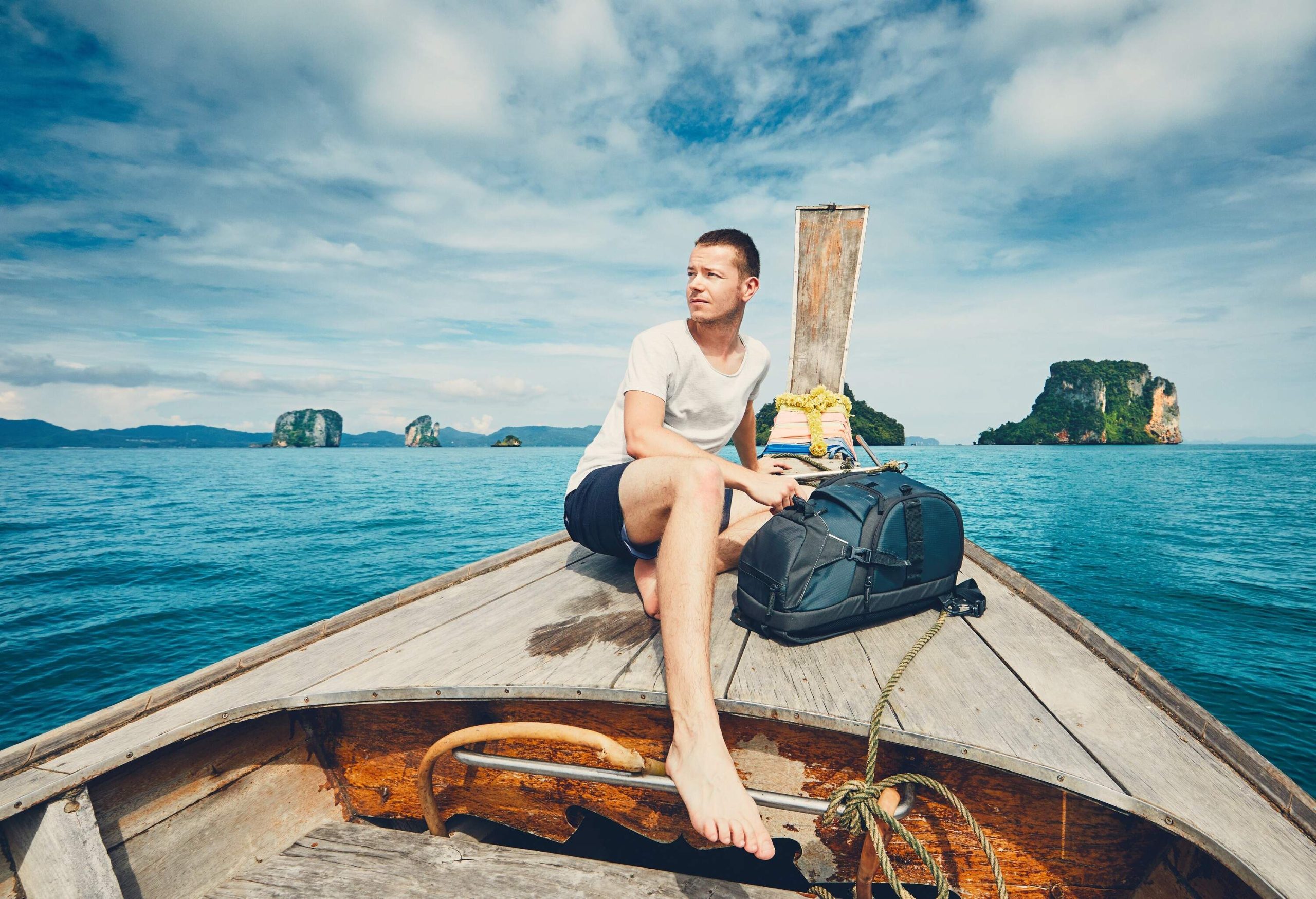 A male traveller sits on the bow of the wooden boat traversing through the enormous protruding rocks on the sea.