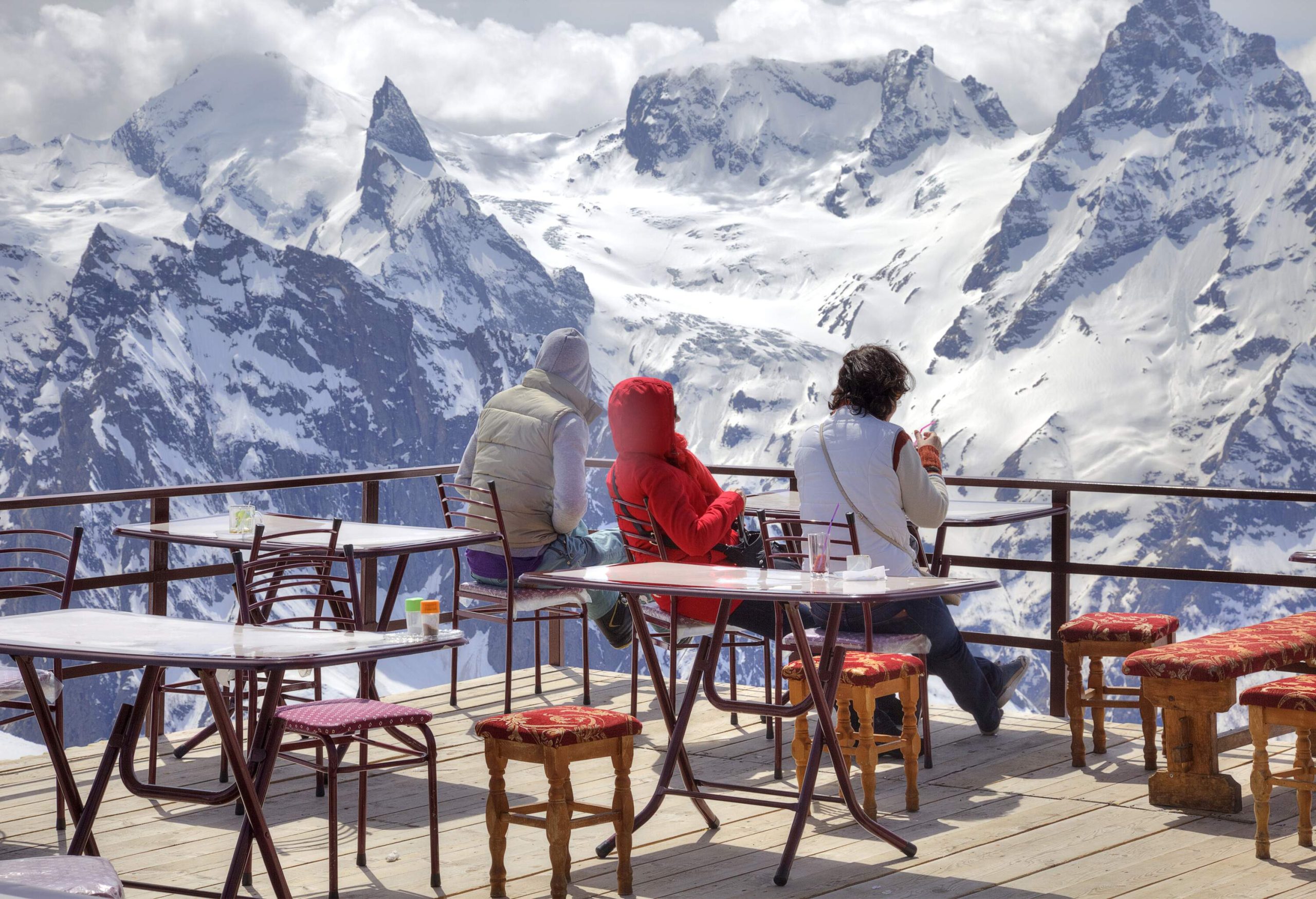 Three people in colourful winter jackets sit on a balcony overlooking the snow-capped mountains.