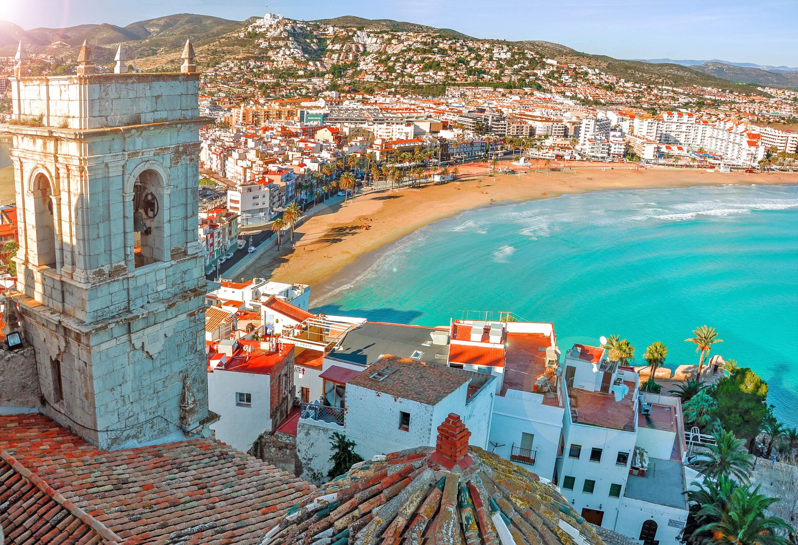 Overlooking a crowded city of tall buildings alongside a pristine beach lined with palm trees viewed from a castle's tower.