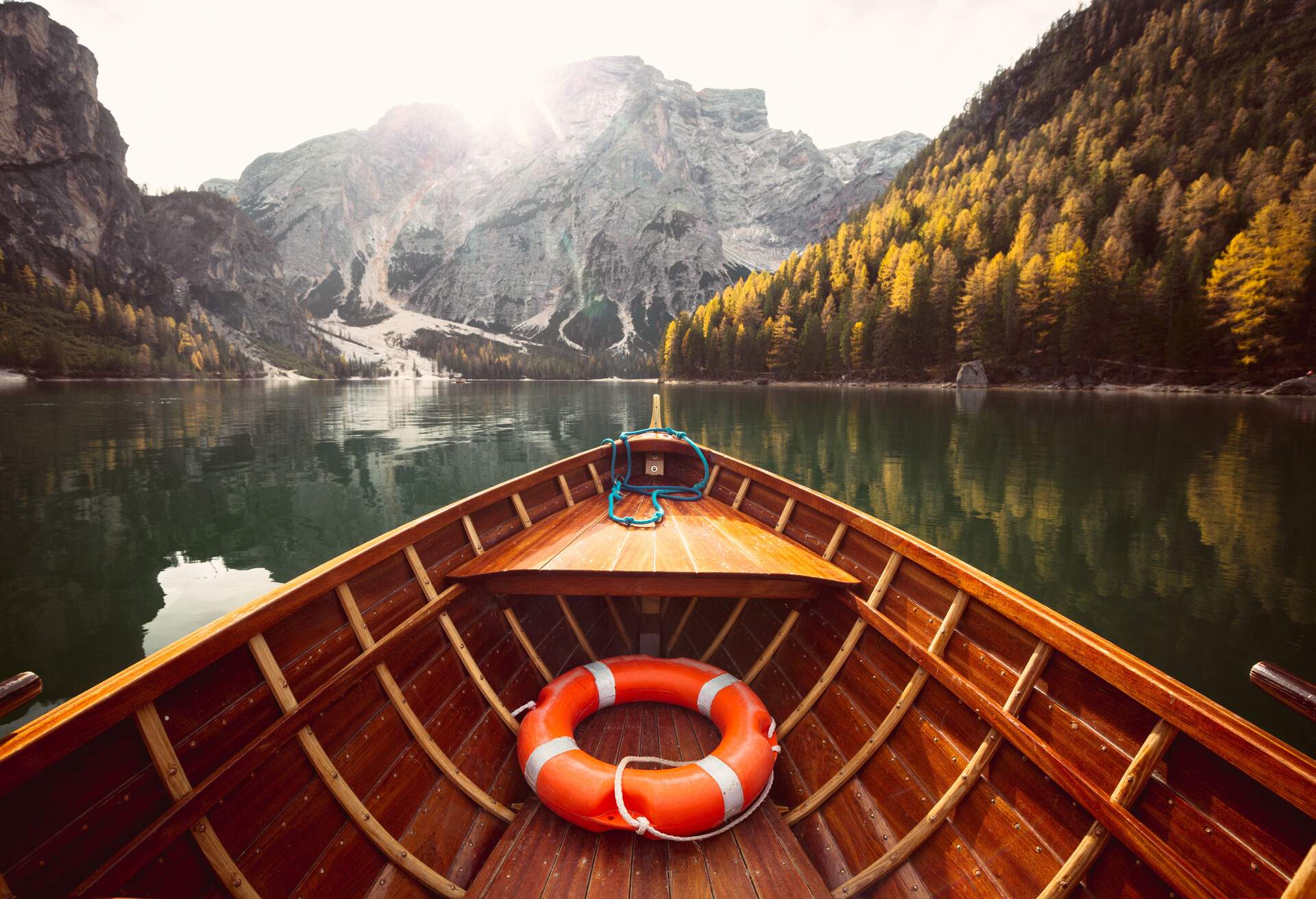A wooden boat with an orange floater on a scenic tranquil lake lined with tall trees in autumn hues beneath the rocky mountains.