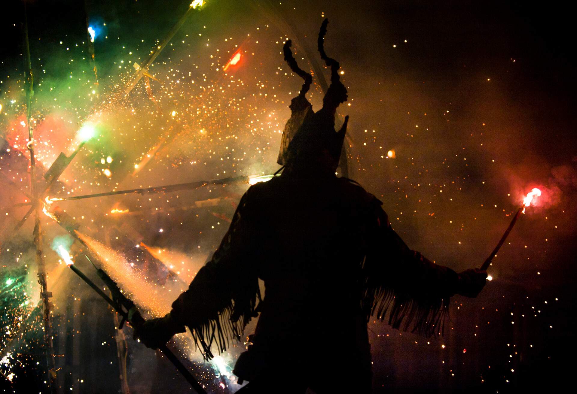 A silhouette of a person in a costume with horns standing in front of the colourful fireworks.