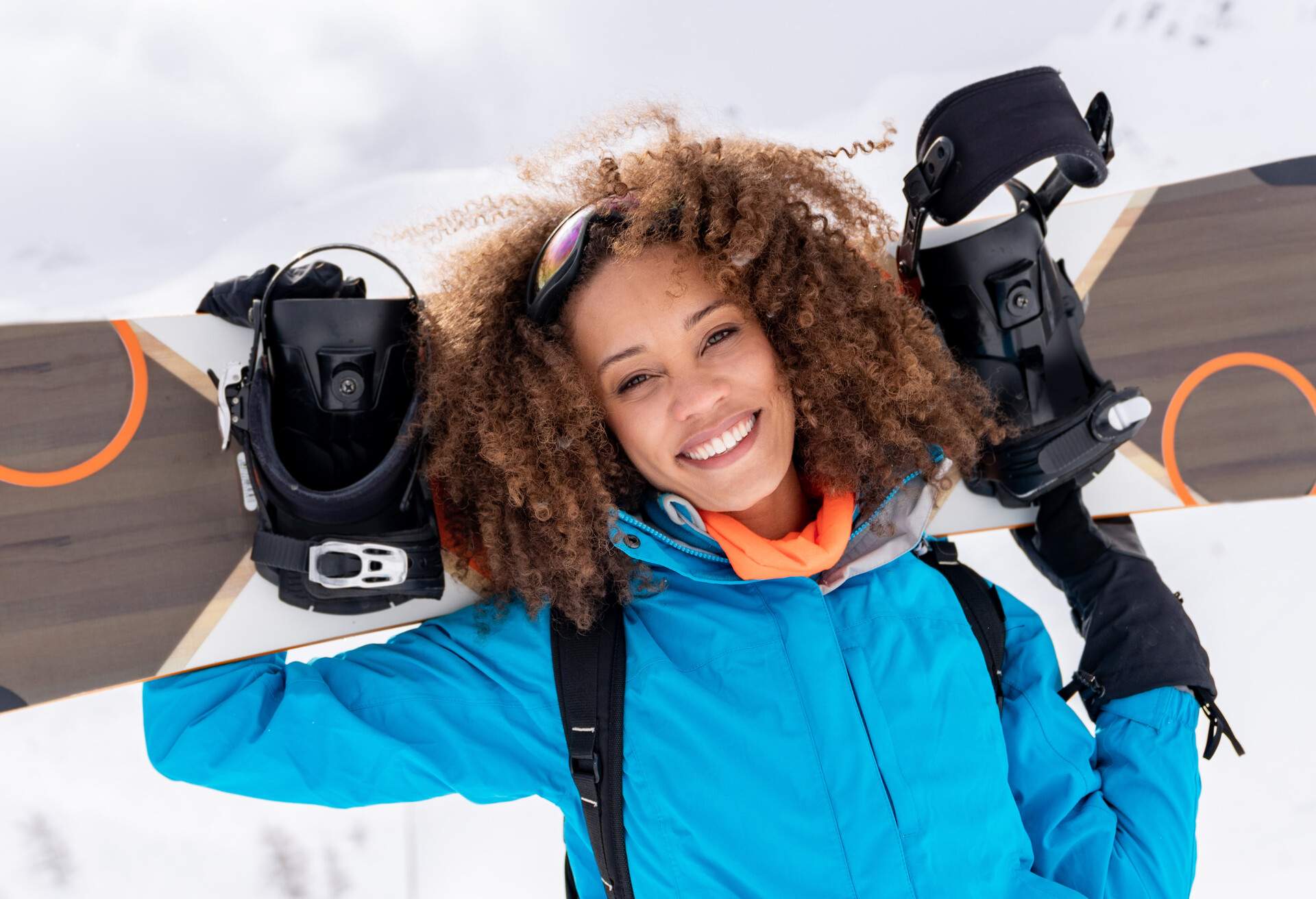 Woman wearing a blue winter shell smiling as she carries a snowboard over her shoulders.