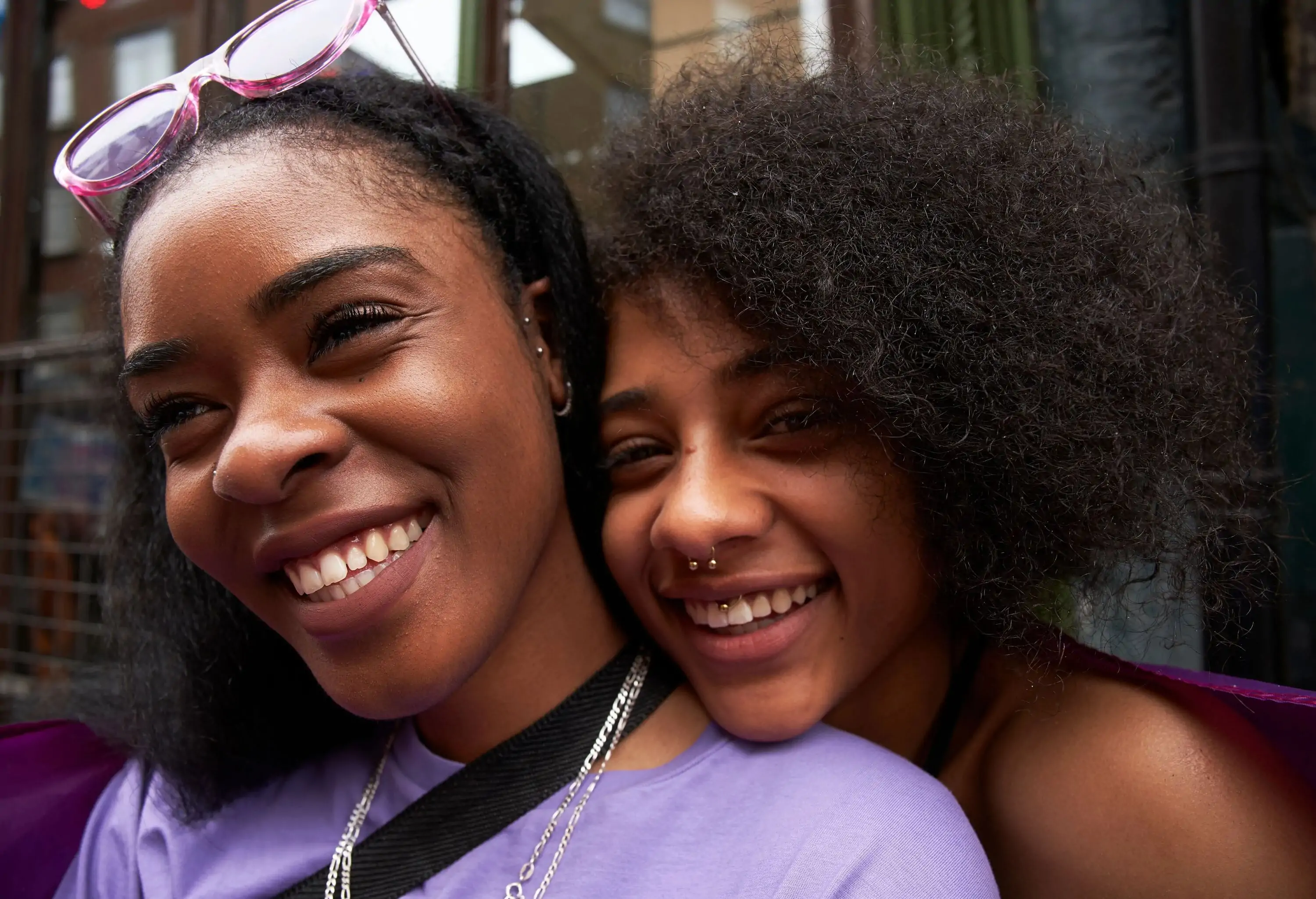 A curly-haired woman smiles as she rests her head on a friend's shoulder.