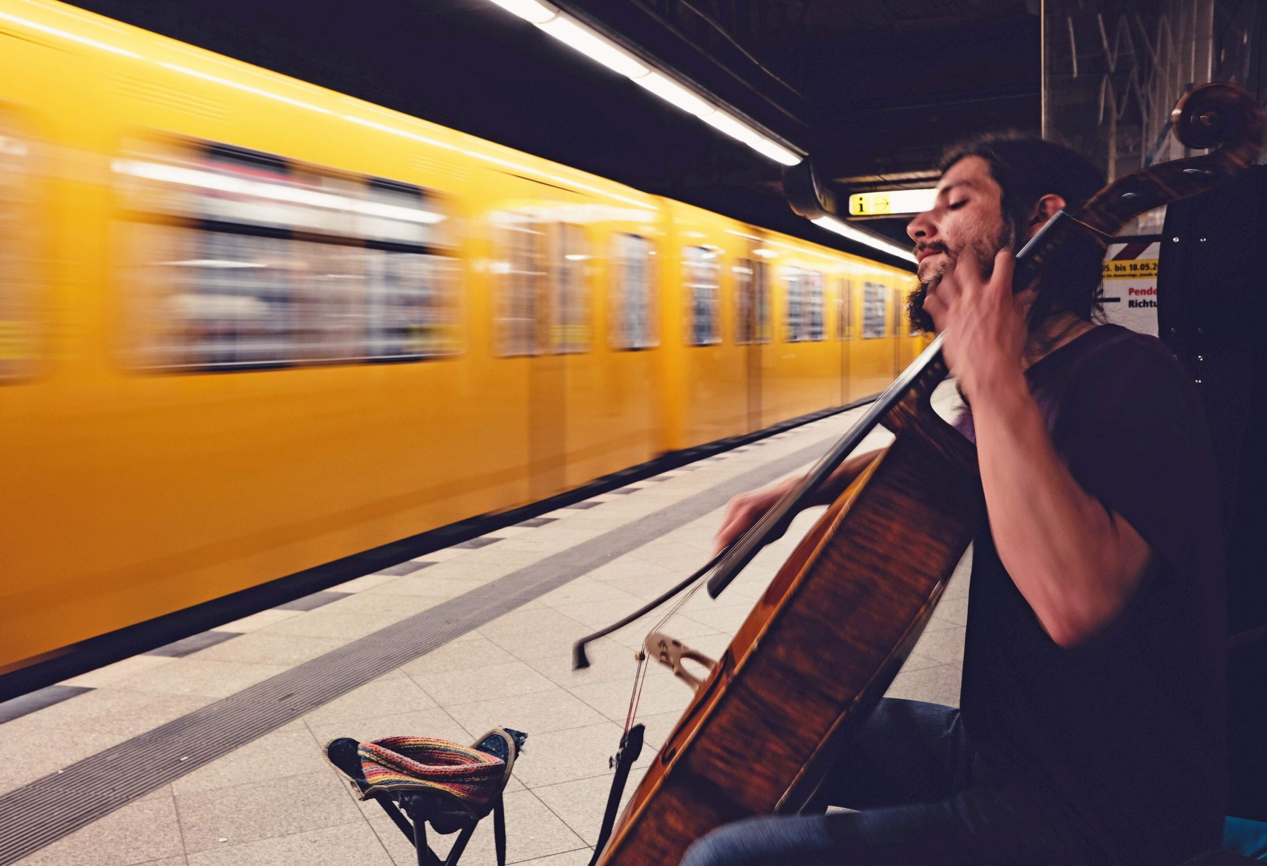 A street performer playing cello in a metro station as a yellow trains goes by. 