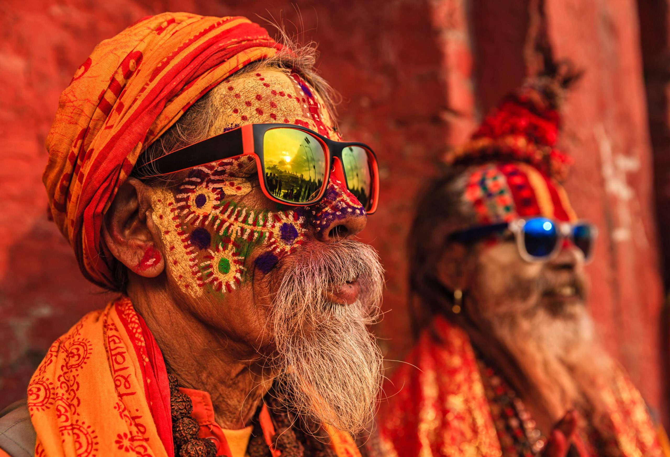 Two sadhus with painted faces wearing sunglasses sit leaning on a wall.