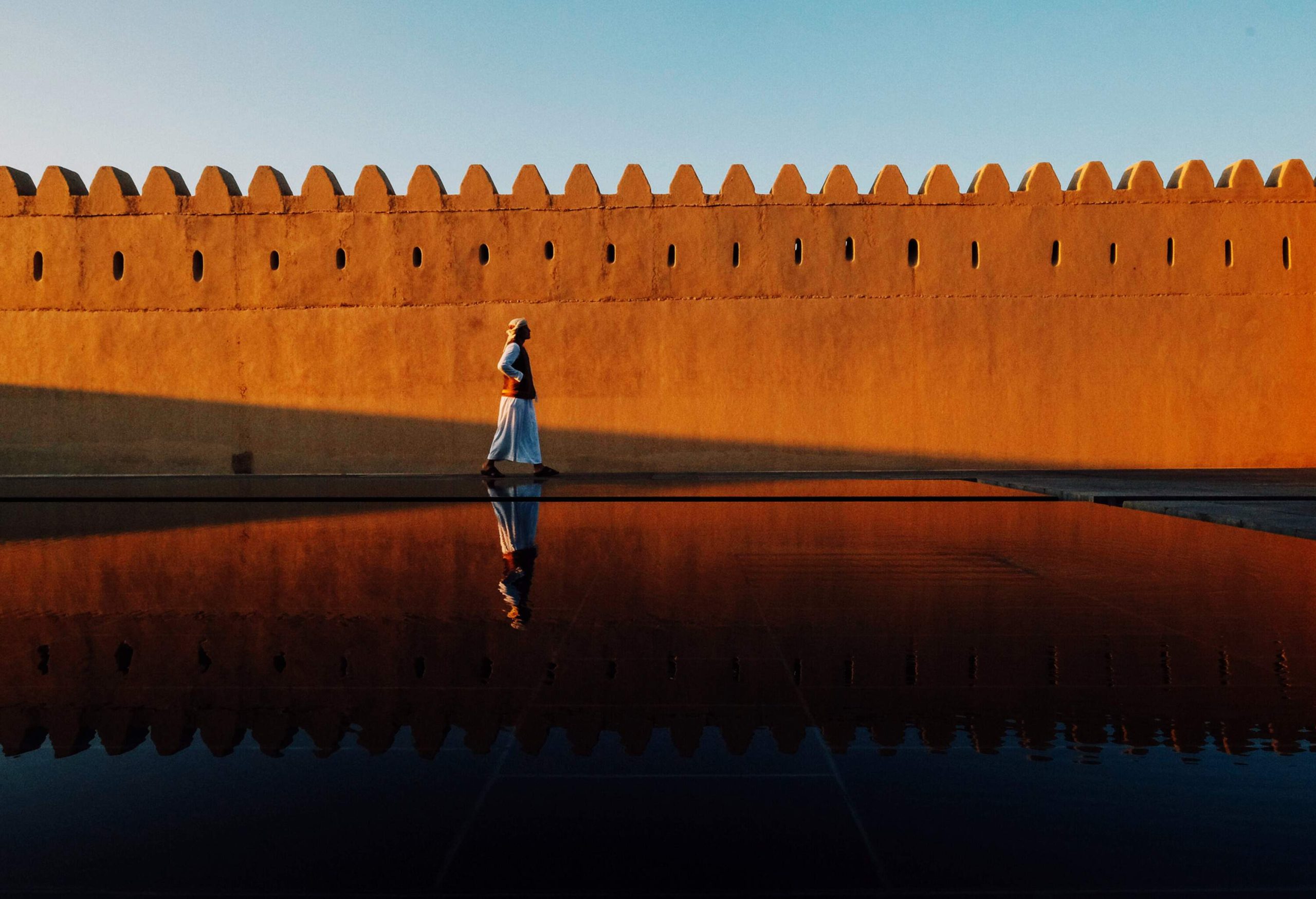 A man walking along a fortress wall with his reflection on the pool of water beside him.