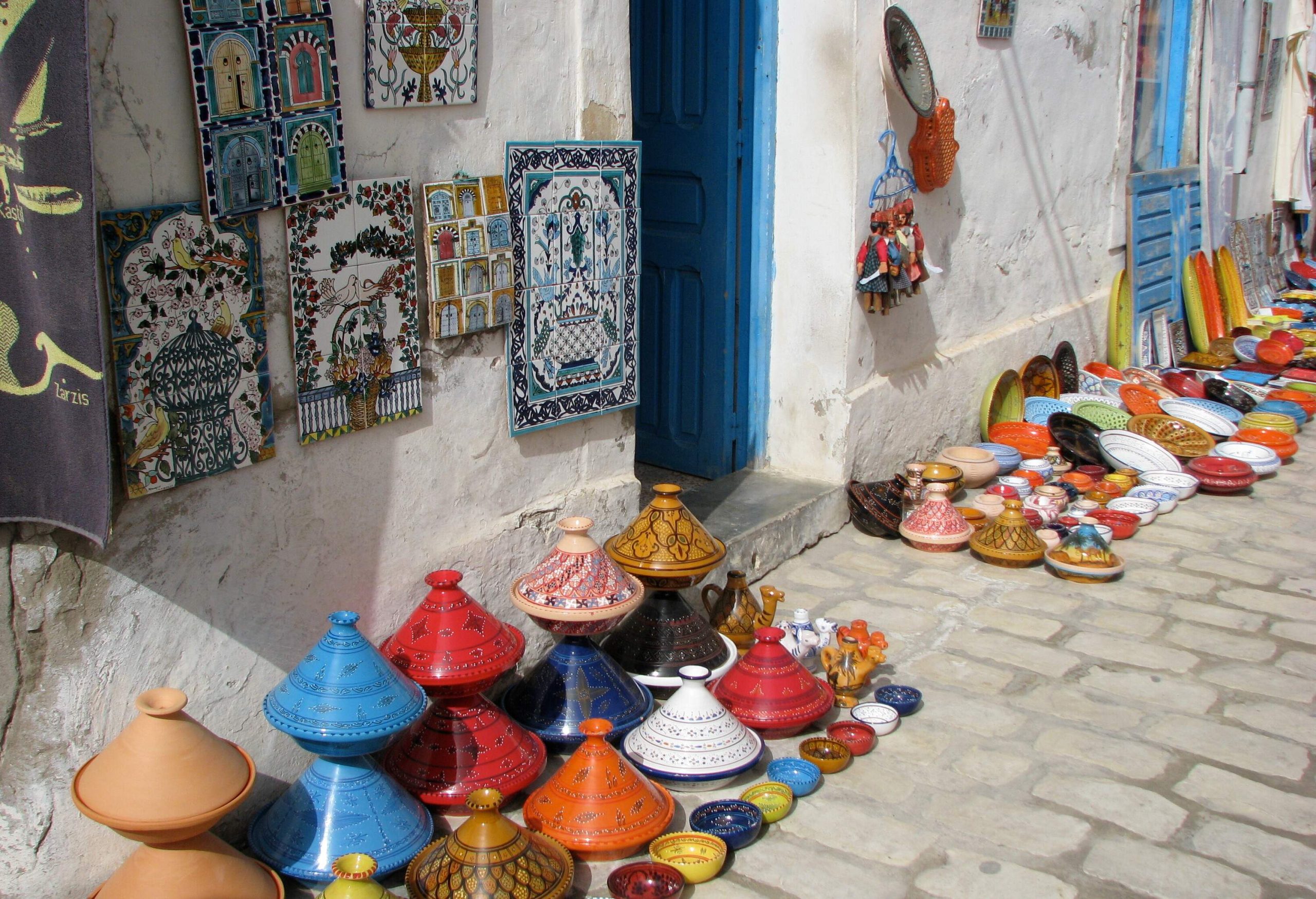 A display of colourful pottery, bowls, and hanged carpets outside a white residence with a blue door.