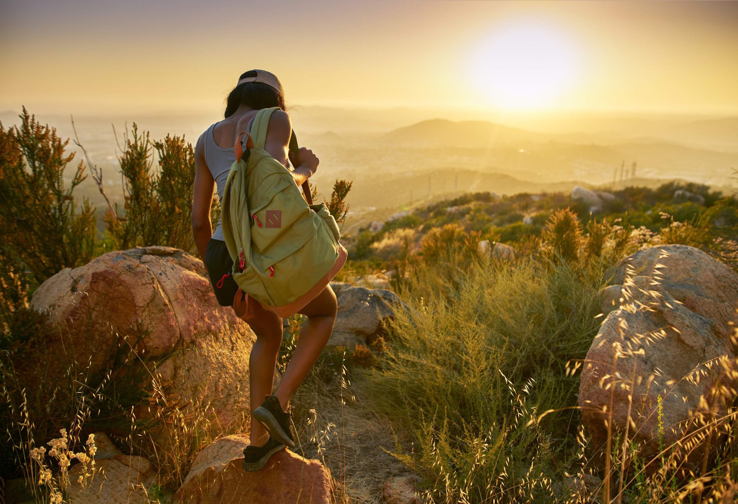 A female hiker with a green rucksack is walking along a rocky path watching the scorching sun over the mountains.
