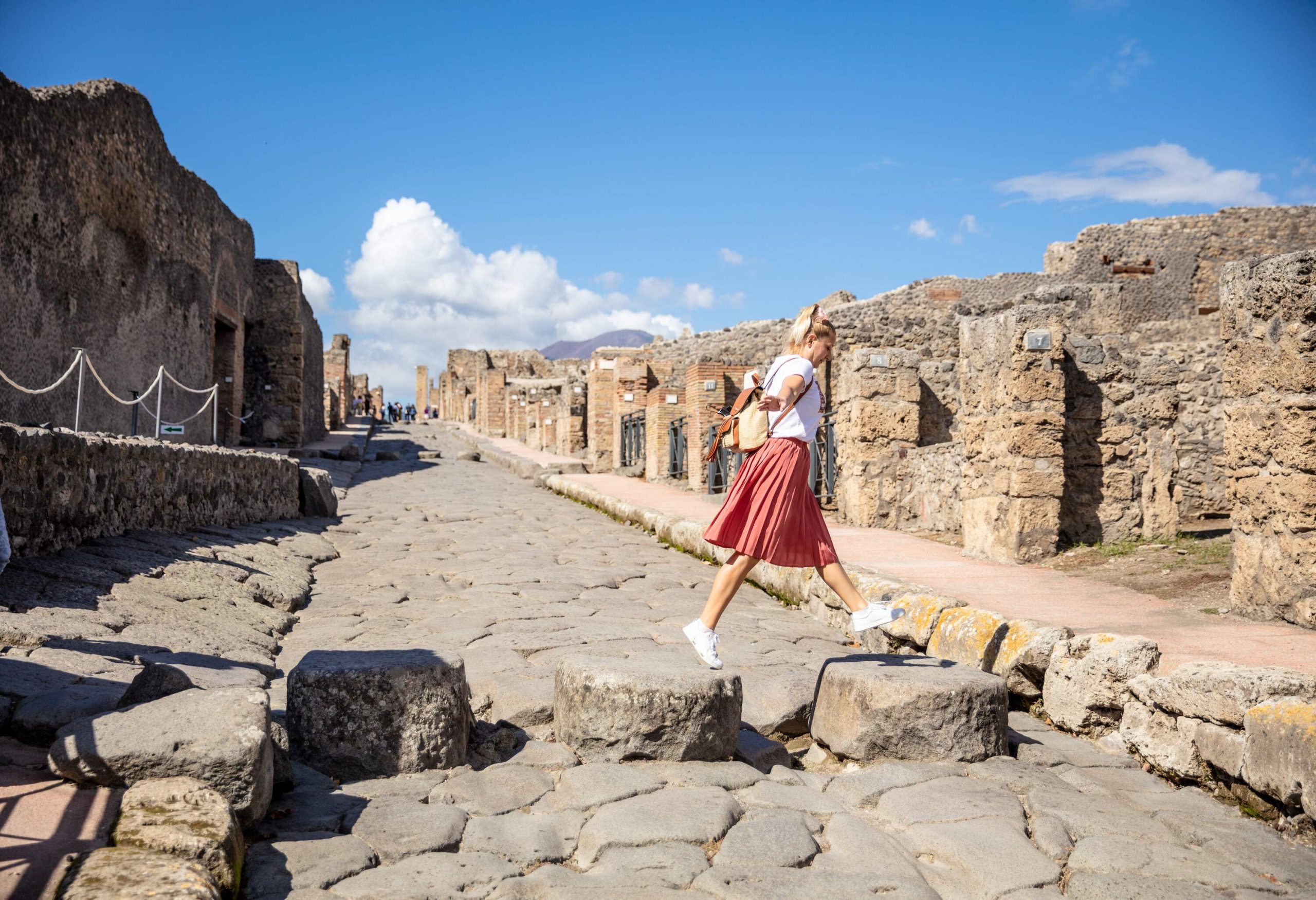 A young blonde lady hops on the rocks in a cobbled street in the middle of the ruins of a vast archaeological site.
