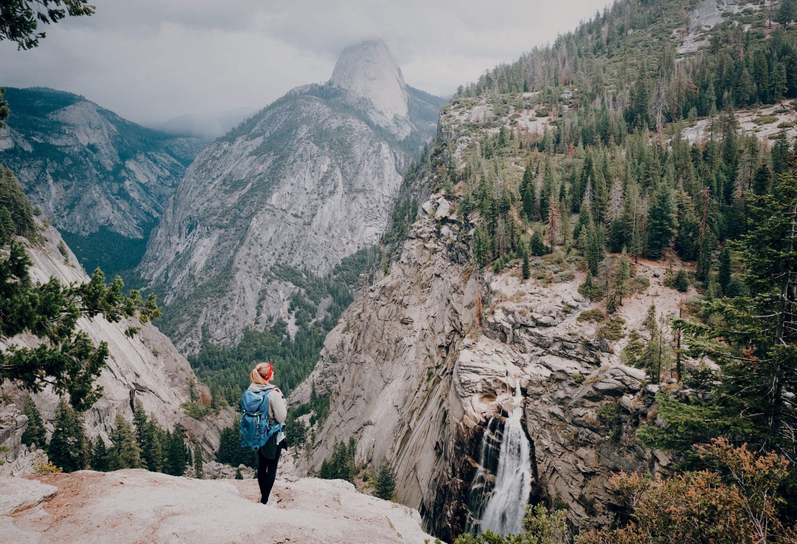 A person with a backpack stands on the edge, gazing at the majestic tree-topped, rugged mountains that lie ahead.