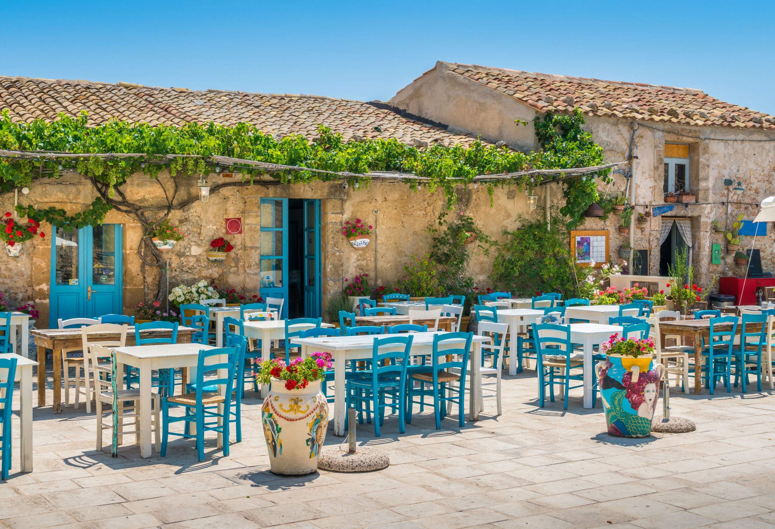 Wooden chairs and tables are set up for al fresco dining outside a charming stone house with a tiled roof, surrounded by lush greenery and a peaceful ambiance.