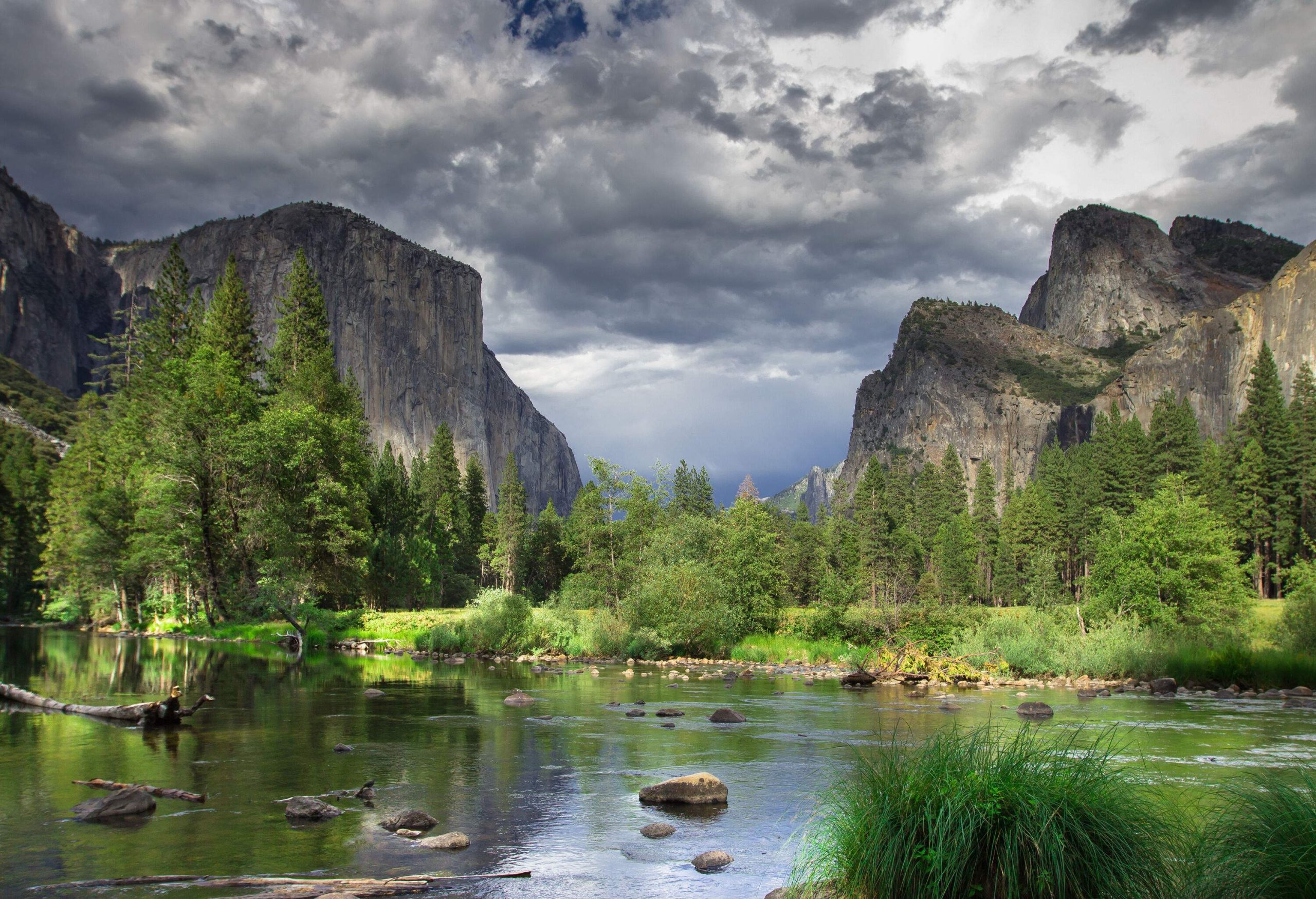 The iconic granite towering rock mountains protrude against an overcast sky along the lush river bank.