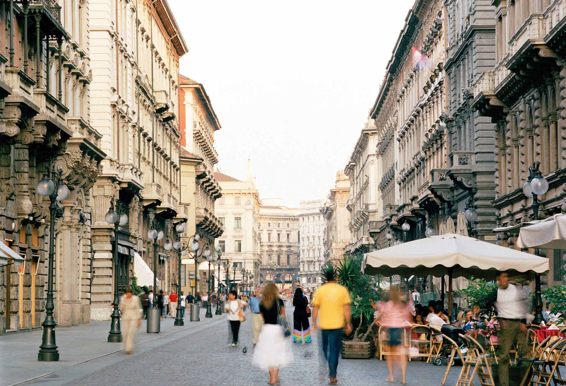 Iron lampposts line the bustling sidewalk, buildings on both sides, and an al fresco restaurant provide a lively scene as people casually wander down this busy street.