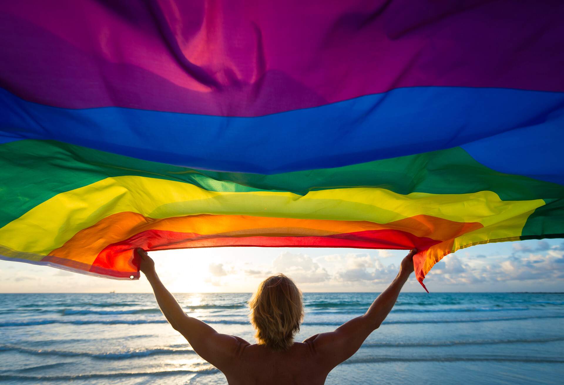 Silhouette of a blonde person raising a rainbow flag blown by the wind in front of a beach.