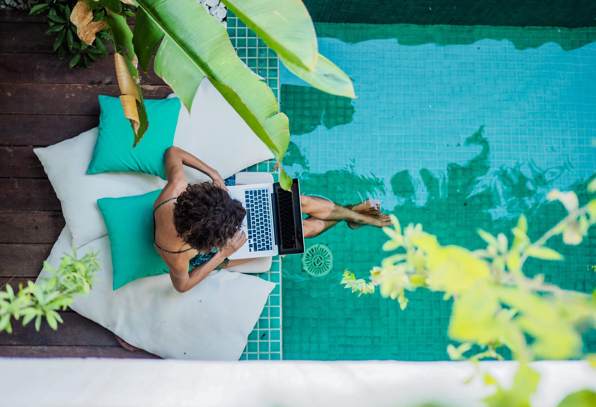 bird view of a remote online working digital nomad women with curly hair and laptop sitting at a sunny turquoise water pool surrounded by cushions and plants in the foreground; Shutterstock ID 1742840084; Purpose: any; Brand (KAYAK, Momondo, Any): any