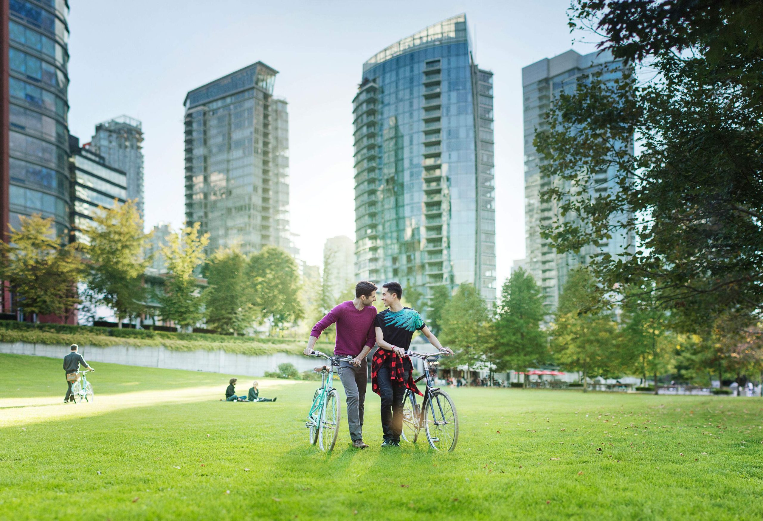 Two cyclists amble side by side, pushing their bicycles through a lush park, as the towering skyscrapers in the background create a striking urban contrast.