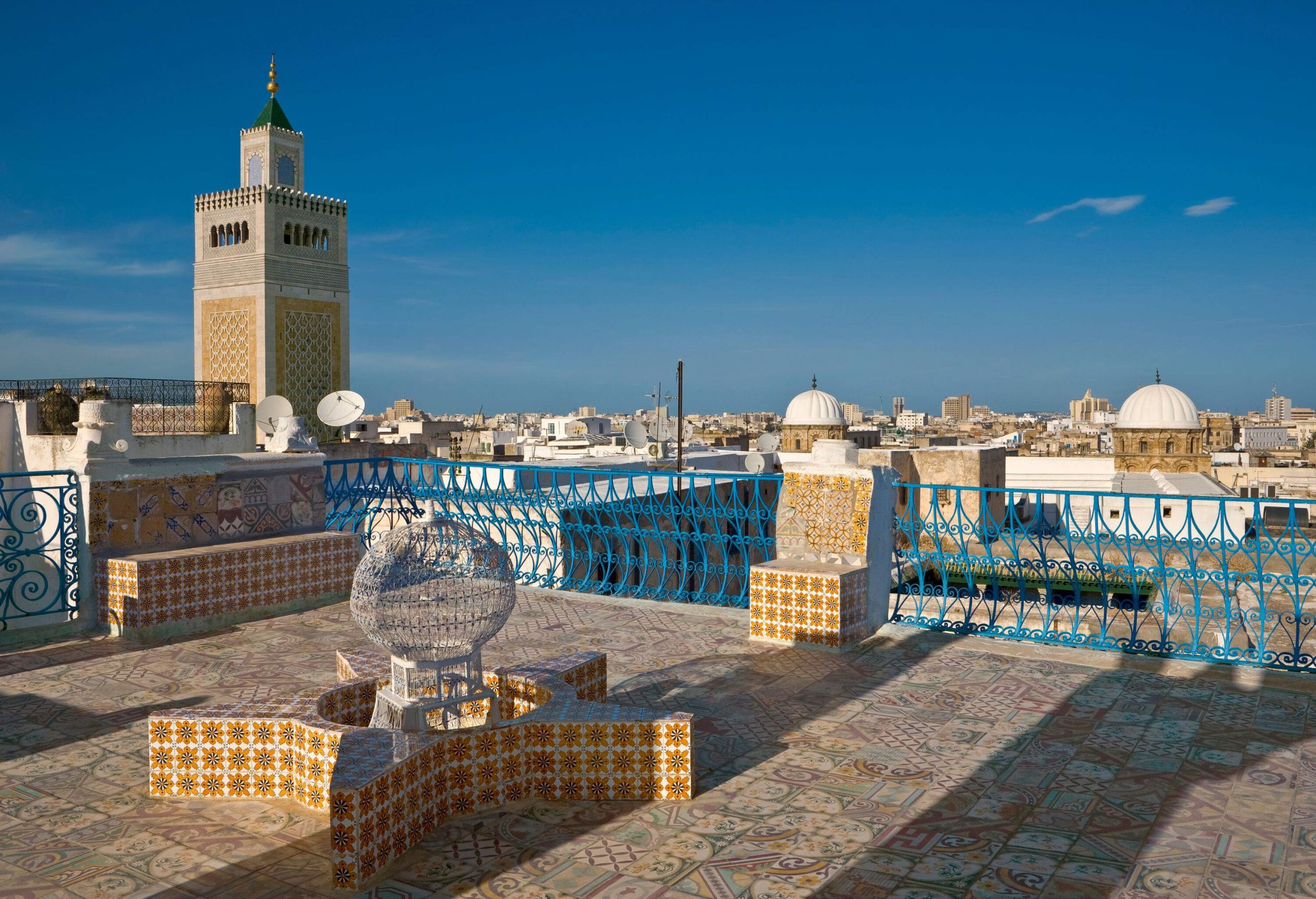 A terrace with intricately ornamented tiles provides a scenic view of an old town skyline, with a mosque minaret standing on the left side.