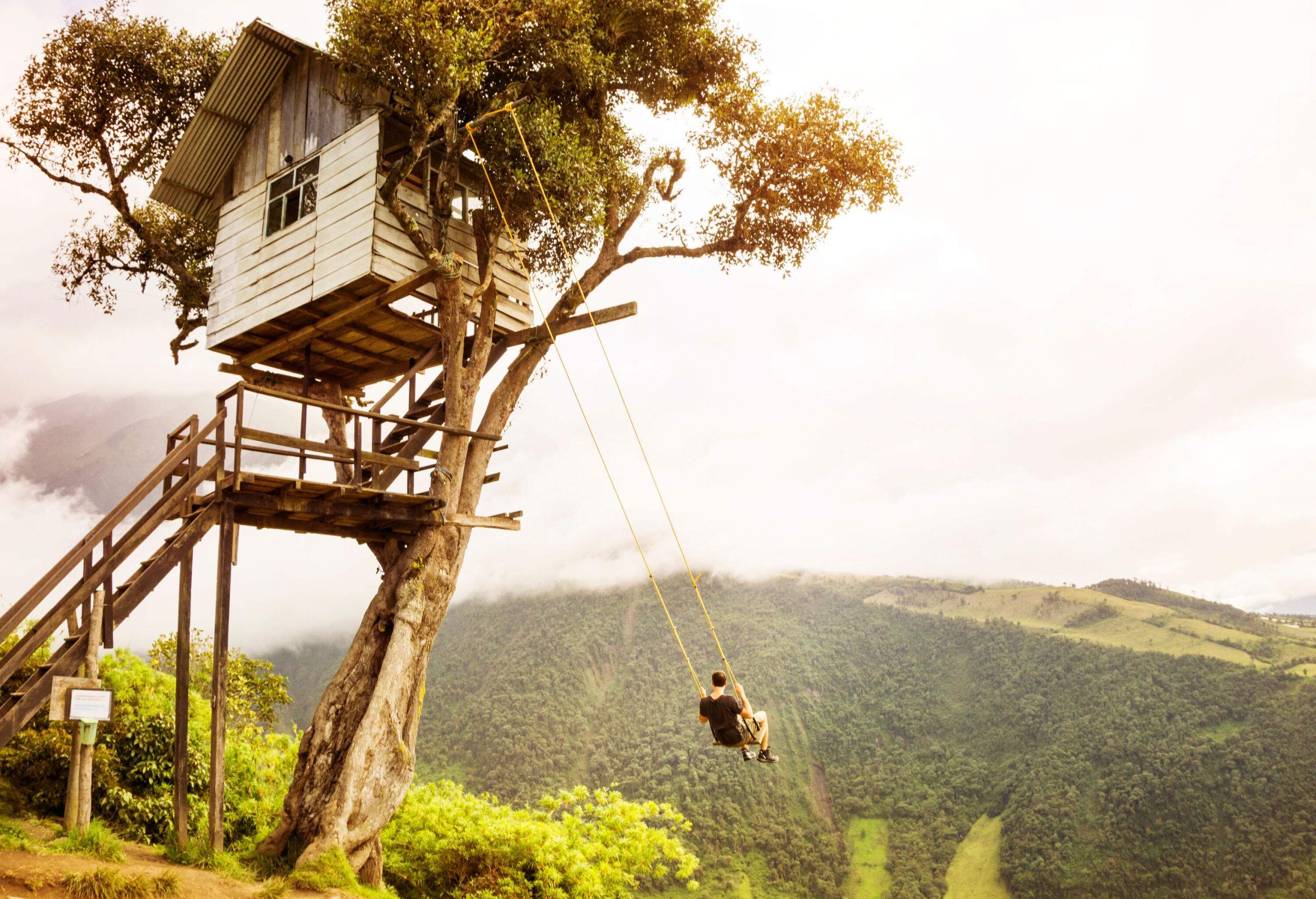 A person on a swing tied to a treehouse perched on a summit, offering a breathtaking view of the majestic forested mountain range under a cloudy sky. 