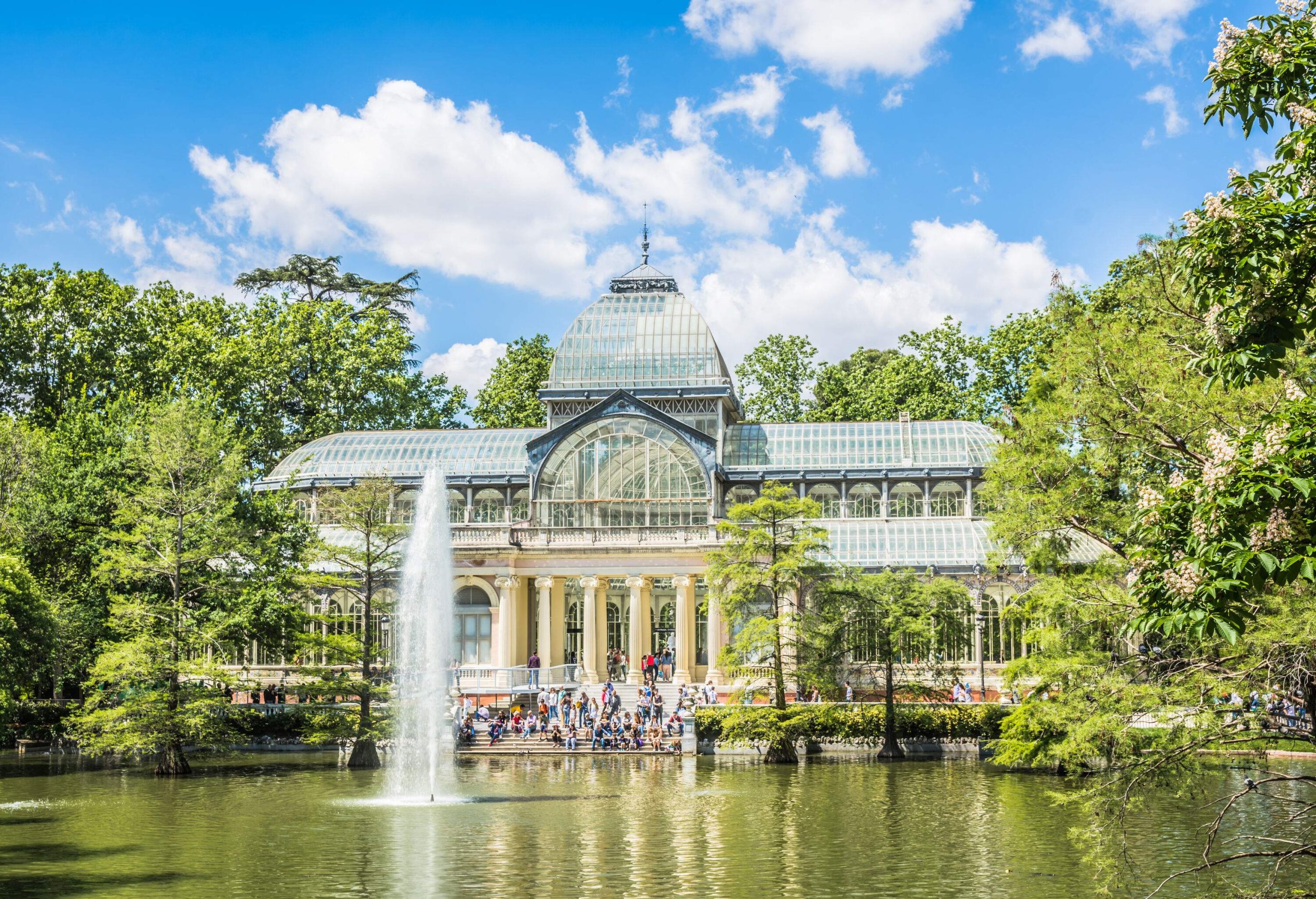 People sitting on the steps outside a glass and steel conservatory overlooking a pond with a water fountain.