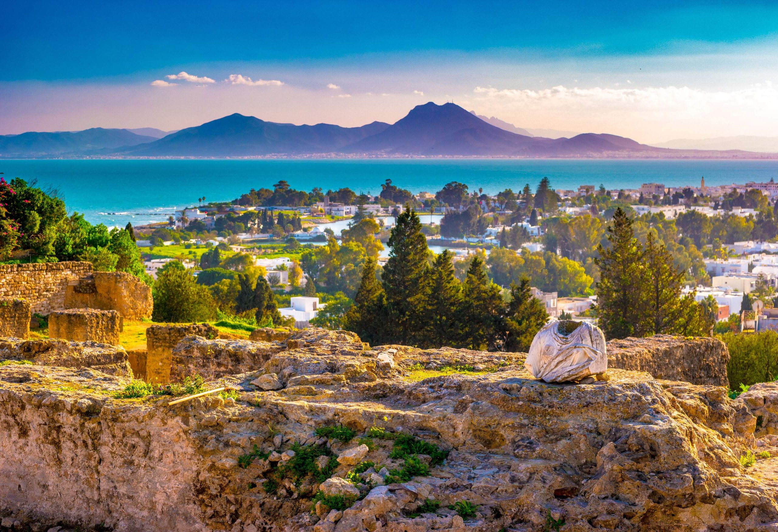 The archaeological site remains of ancient Carthage, perched on a hill overlooking a coastal town, with the sea surrounding it and the silhouette of a majestic mountain range in the distance.