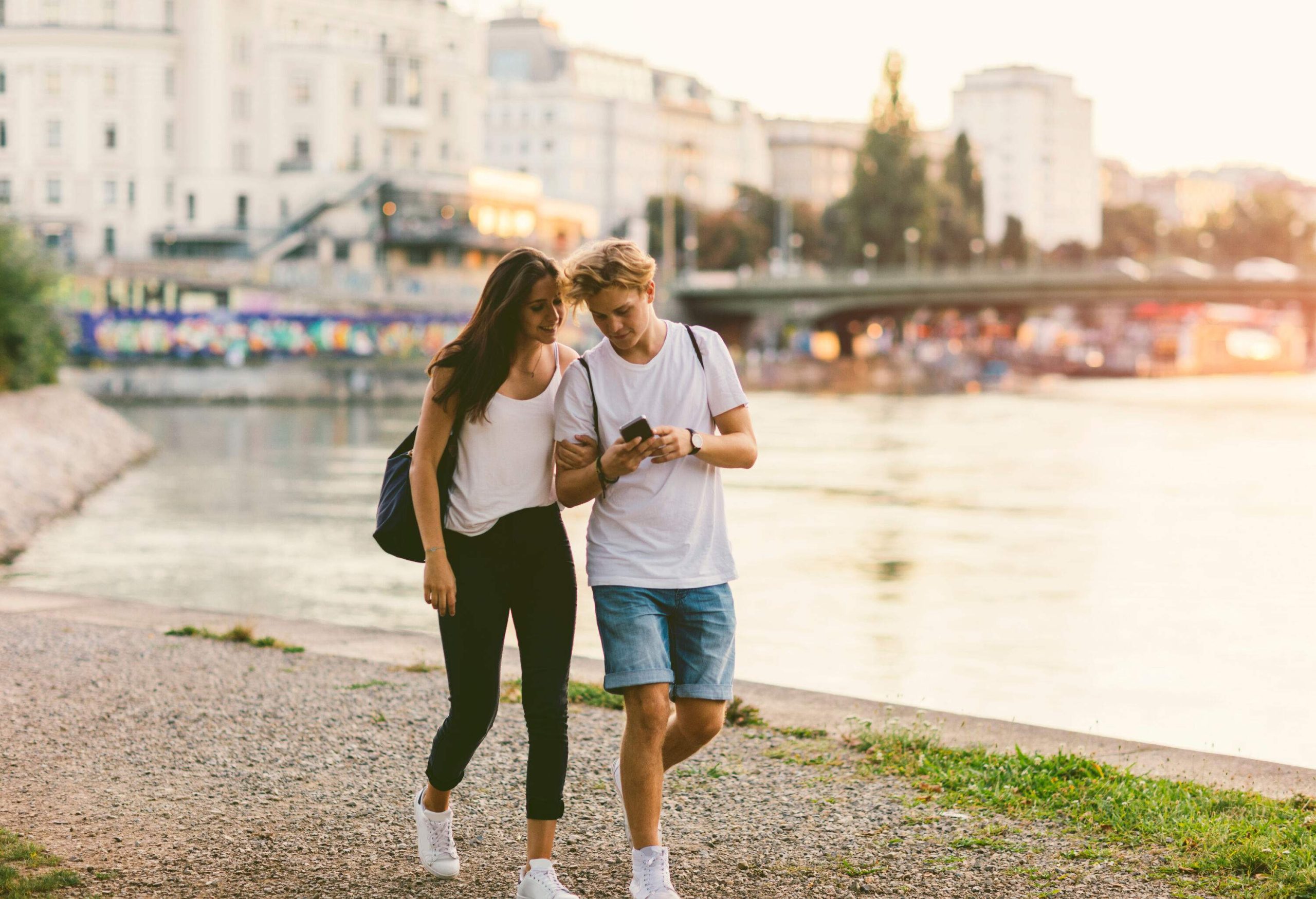 A teenage couple strolls leisurely by the Danube River, the boy holding a smartphone and sharing its content with his companion, both smiling and dressed in casual attire.