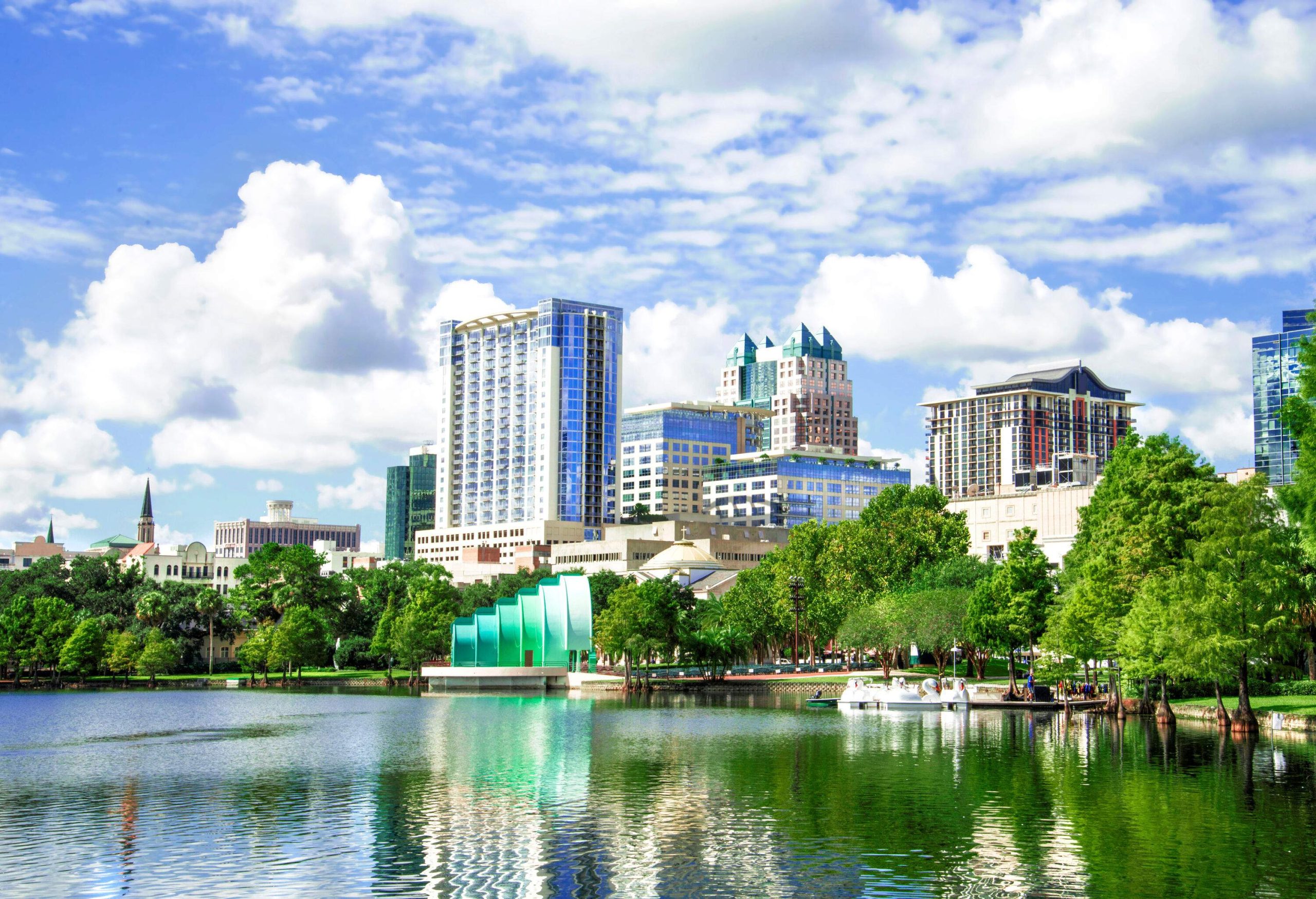 A tranquil lake lined with tall green trees with a view of modern urban cityscape against the cloudy blue sky.