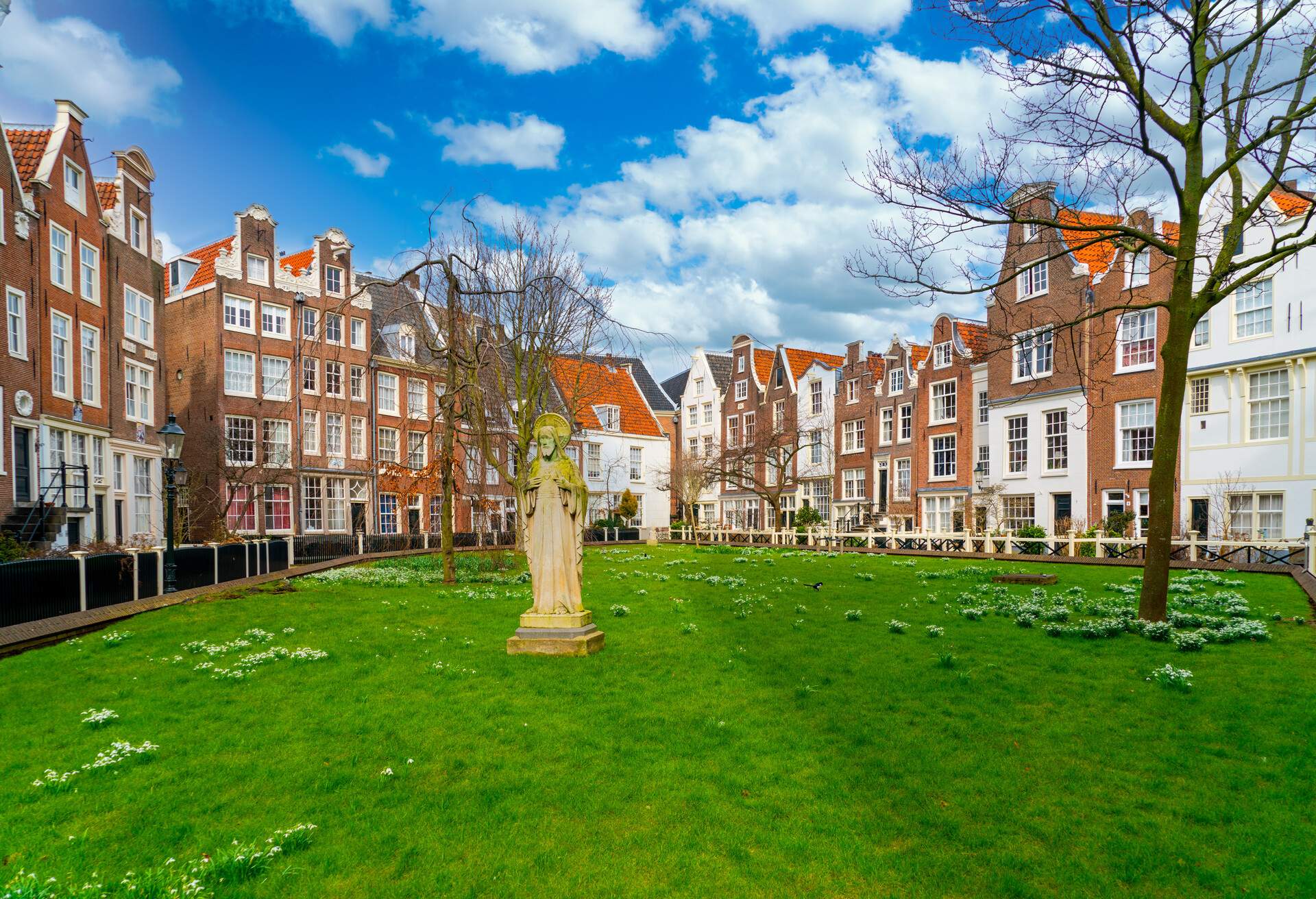 A statue in the middle of a park in Amsterdam surrounded by narrow buildings and with green grass and spring flower on the ground