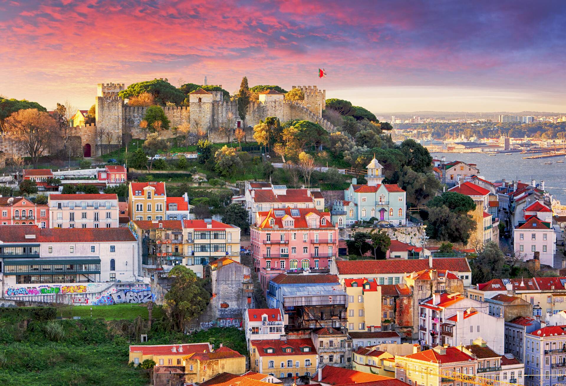 São Jorge Castle is a fortified castle perched on a forested hill surrounded by colourful buildings in a coastal city.