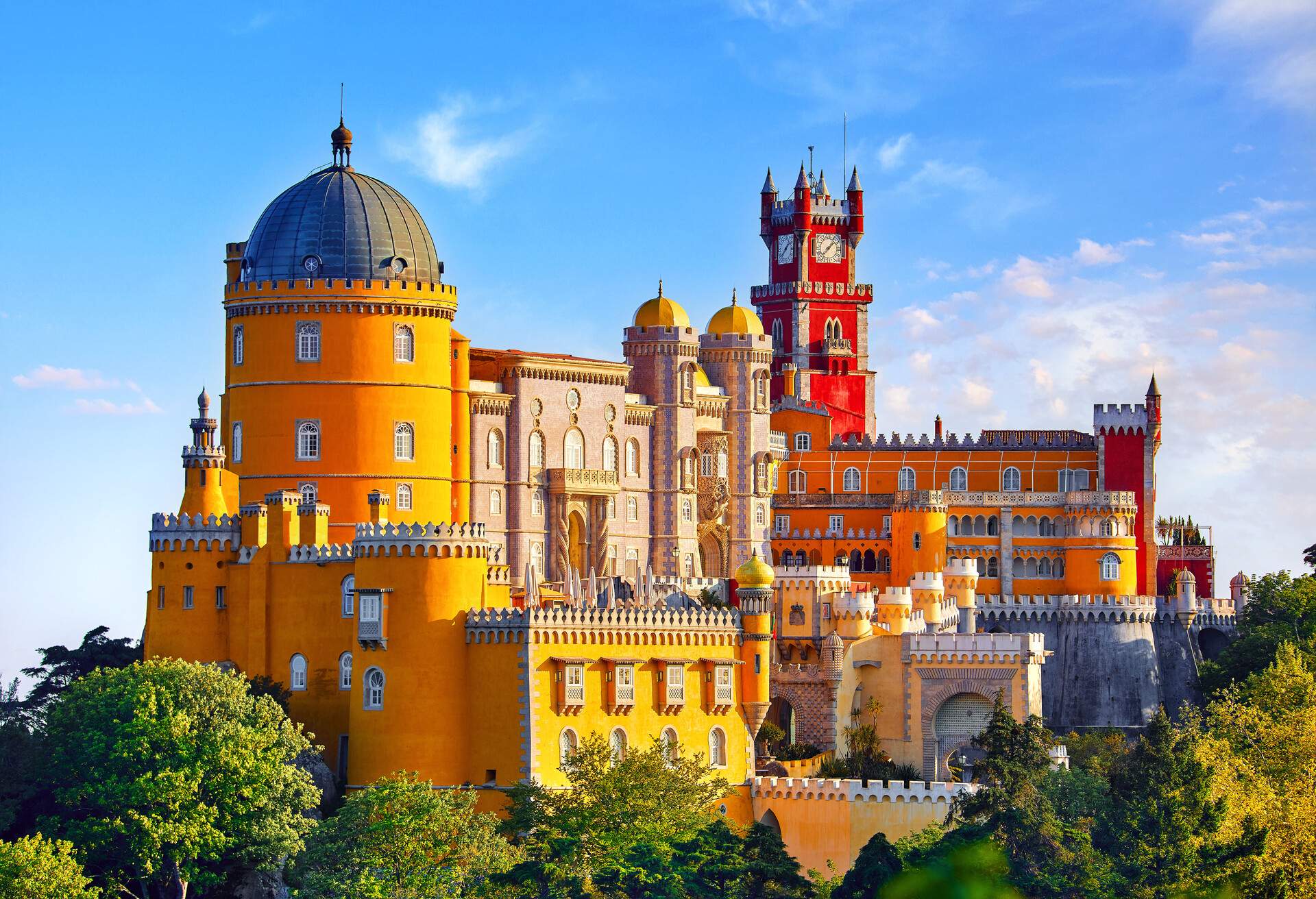 Pena Palace is a vibrant and colourful hilltop castle in yellow and red hues with domed towers surrounded by crenellated structures and a clock tower with turrets.