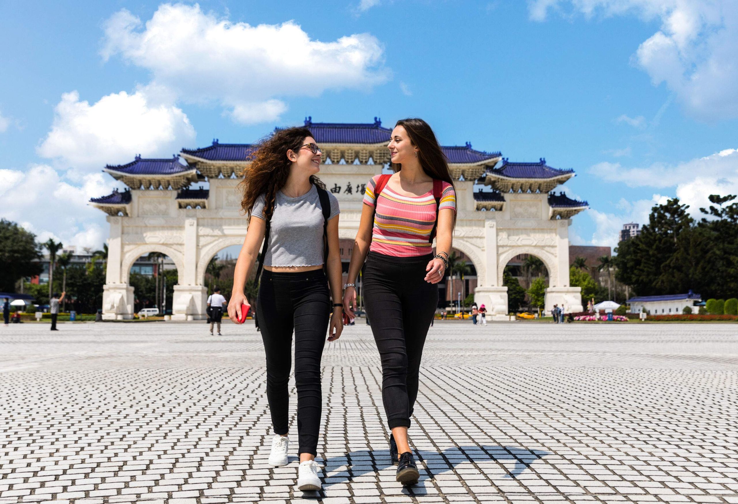 Two young women walking across a paved public plaza in front of a Chinese-style gateway structure.