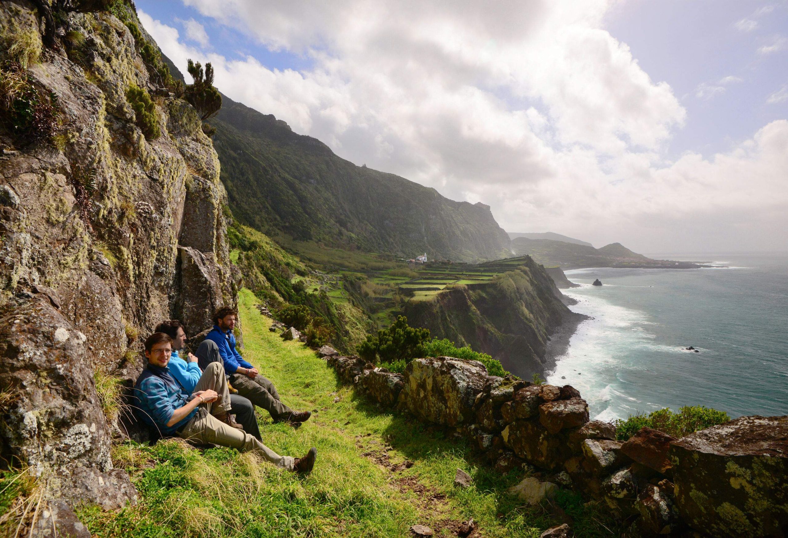 Three individual leans on the rock wall of a lush island cliff overlooking the sea against the cloudy sky.