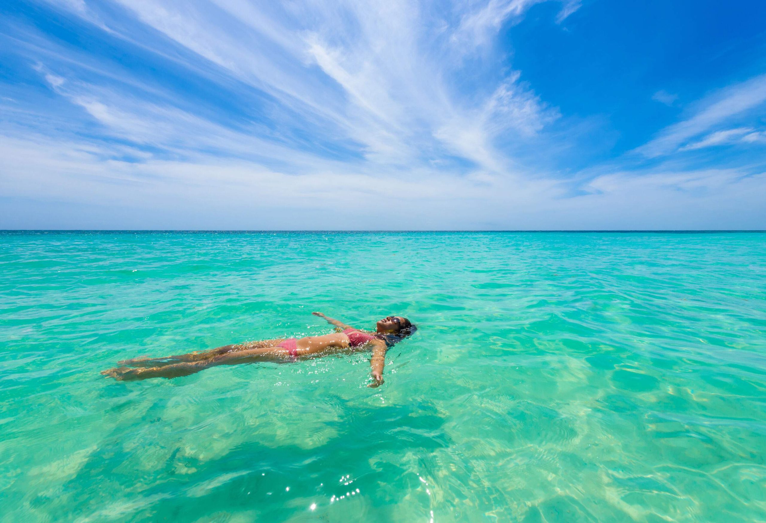 A carefree young woman in a bikini blissfully relaxes in the vibrant turquoise waters, effortlessly back-floating amidst the seemingly endless expanse of the vast sea.