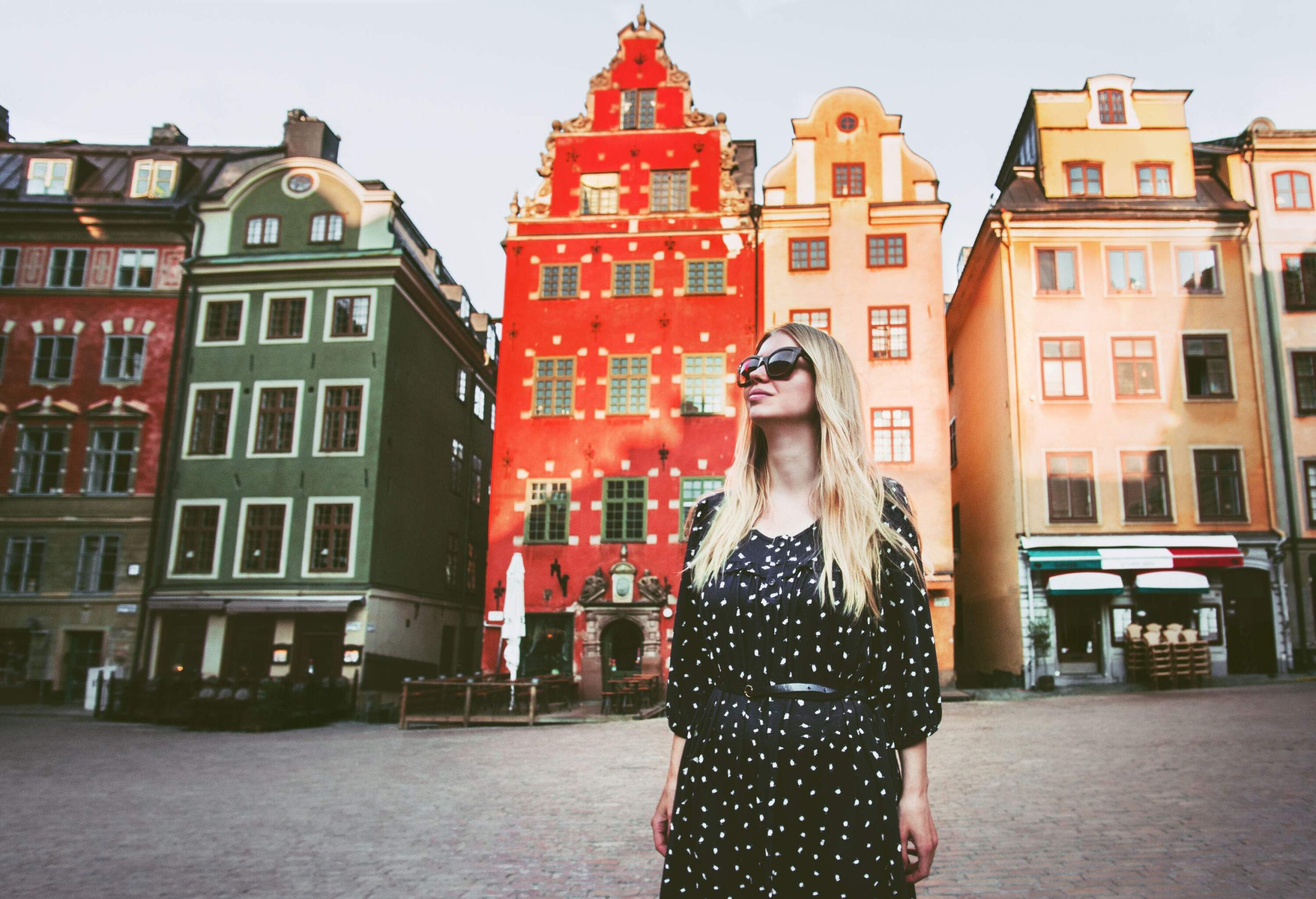 A blonde woman in a black dress and sunglasses stands in the middle of a public square surrounded by colourful buildings.