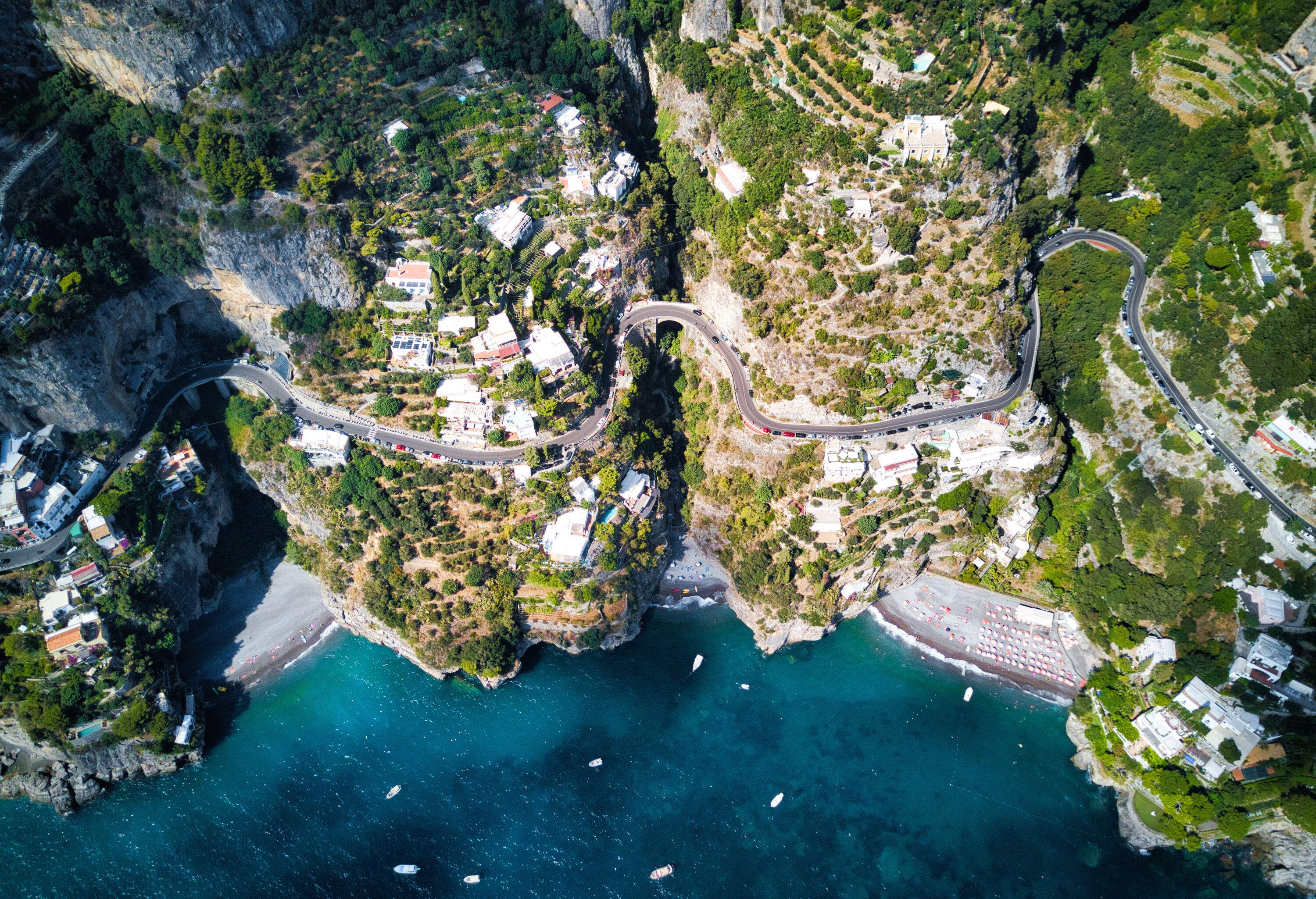 A winding road across a hillside coastal town with terraced buildings and lush trees.