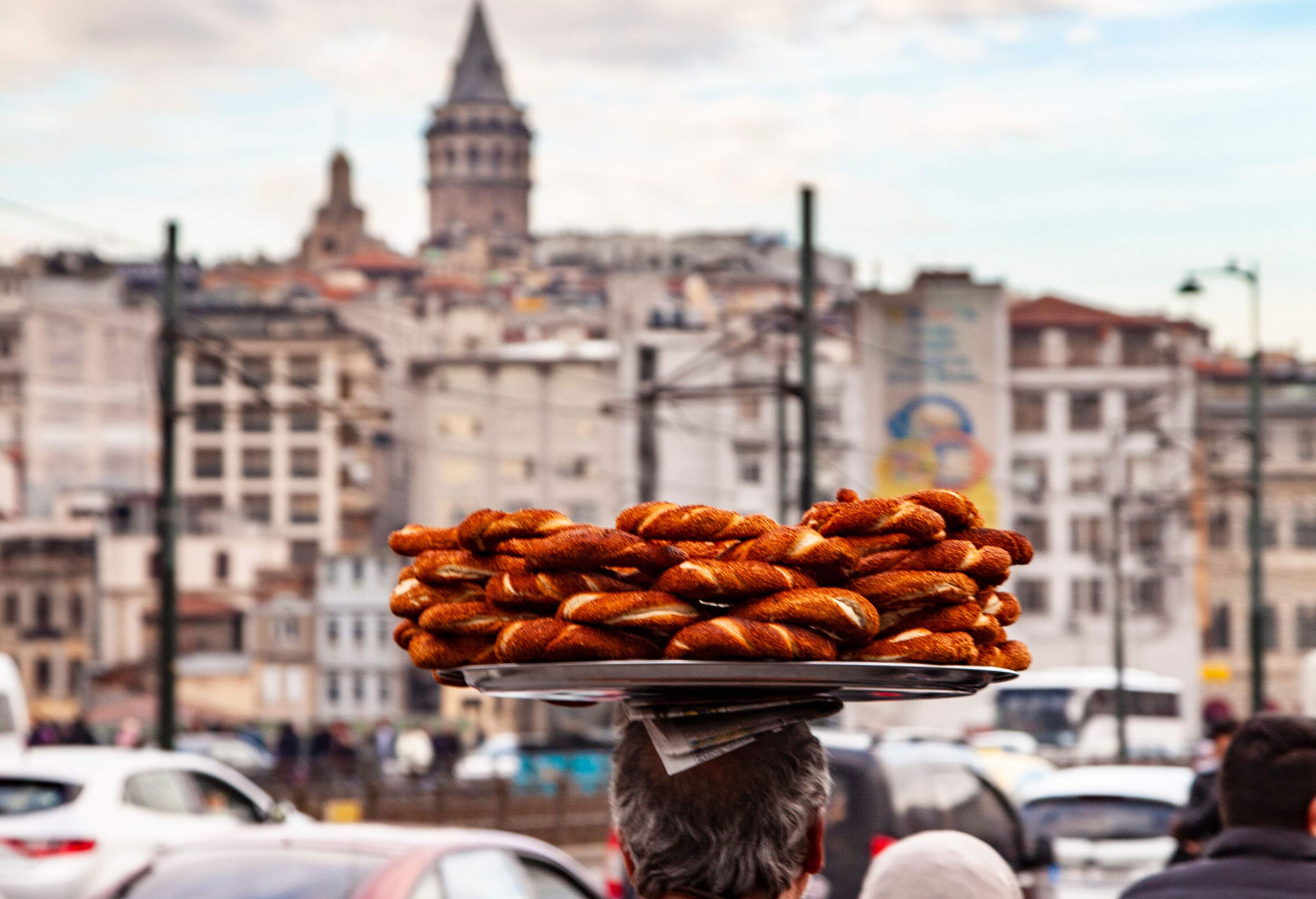 A man skillfully balances a large tray of pastries on his head as he confidently walks along a bustling pedestrian road. 